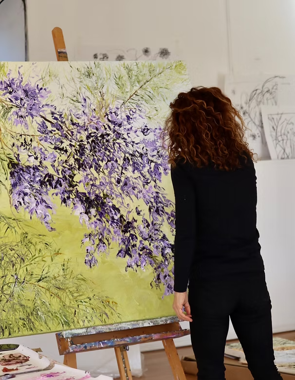 Woman with curly red hair painting or viewing a large canvas with purple and green floral artwork in an art studio.