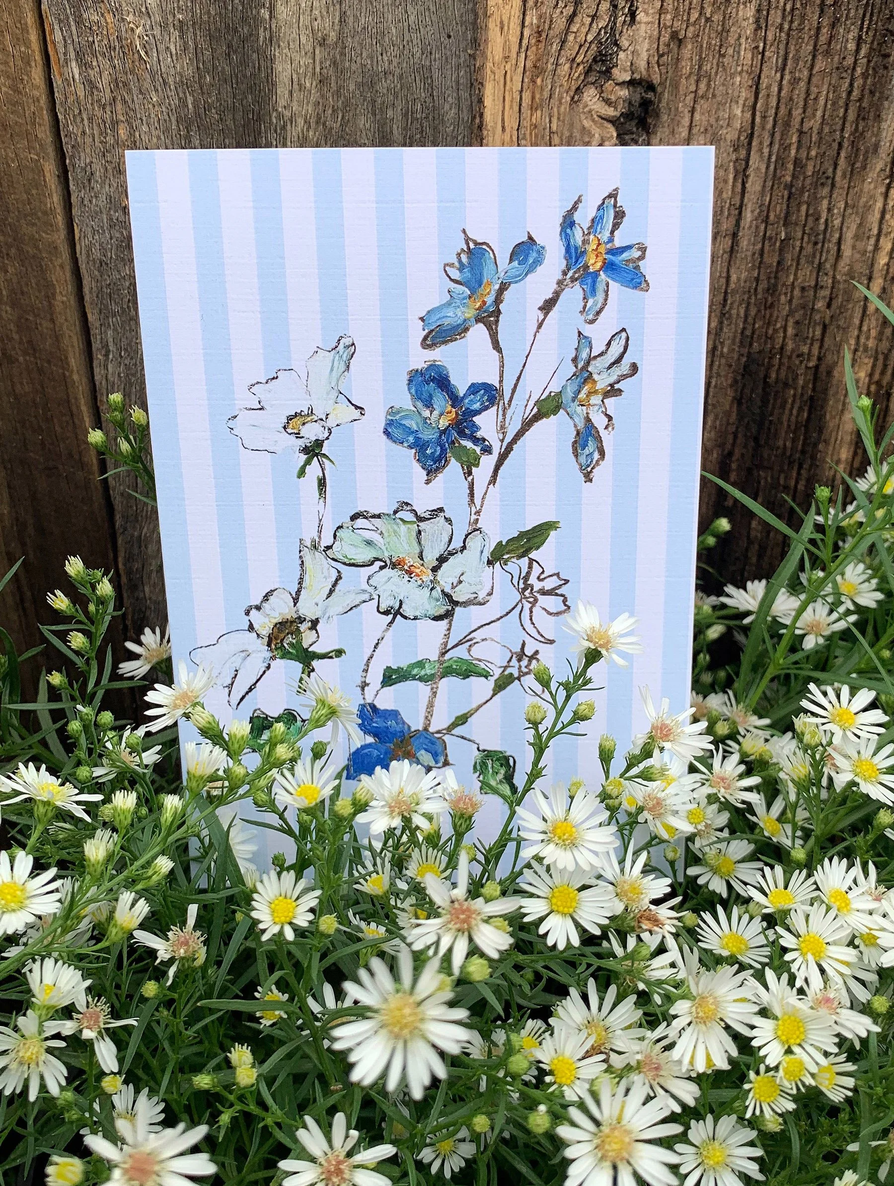 A floral illustration on lined paper featuring blue and white flowers, set against a rustic wooden background and surrounded by a garden of small white daisies with yellow centers.