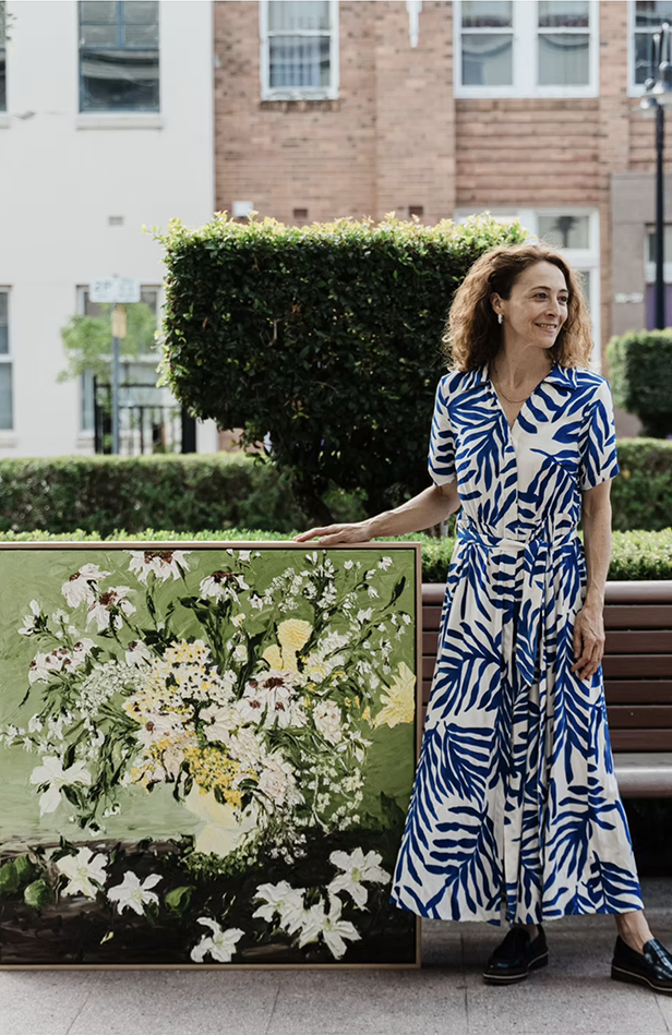 A woman in a blue and white patterned dress stands outdoors next to a large floral painting. She has curly hair and is smiling, with a background of trees, bushes, and residential buildings.