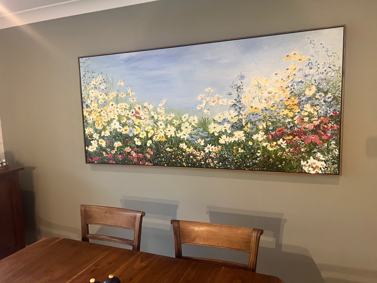 A large painting of a colorful flower garden with blue sky, hanging on a light green wall in a dining room with wooden table and chairs.