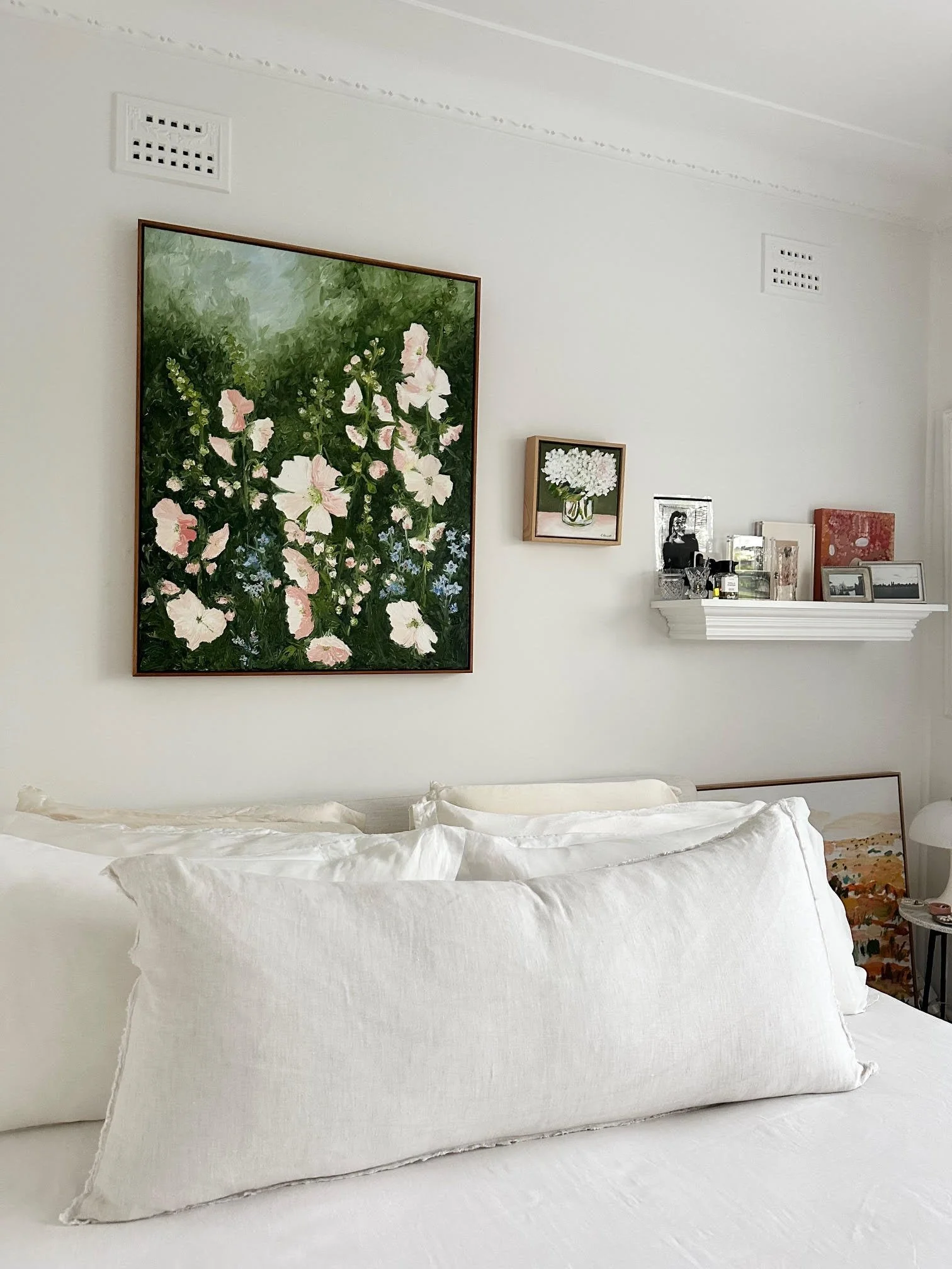 A bedroom with a white bed featuring large pillows, a white wall with three floral art pieces including one large floral painting and two smaller framed photos, and a white shelf with decorative items.