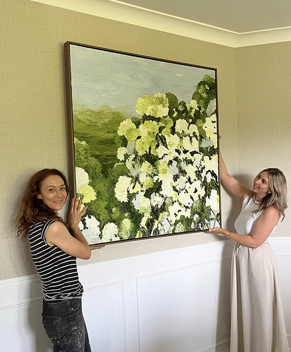 Two women standing in front of a large painting of green and white flowers on a wall in a room with beige walls and white wainscoting.