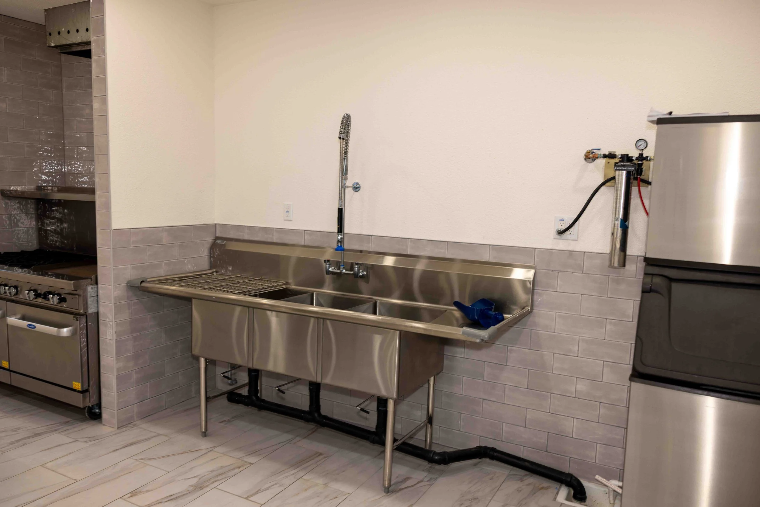 Commercial stainless steel kitchen sink with a flexible hose faucet, gray tiled wall, and nearby appliances in a clean, industrial kitchen setting.