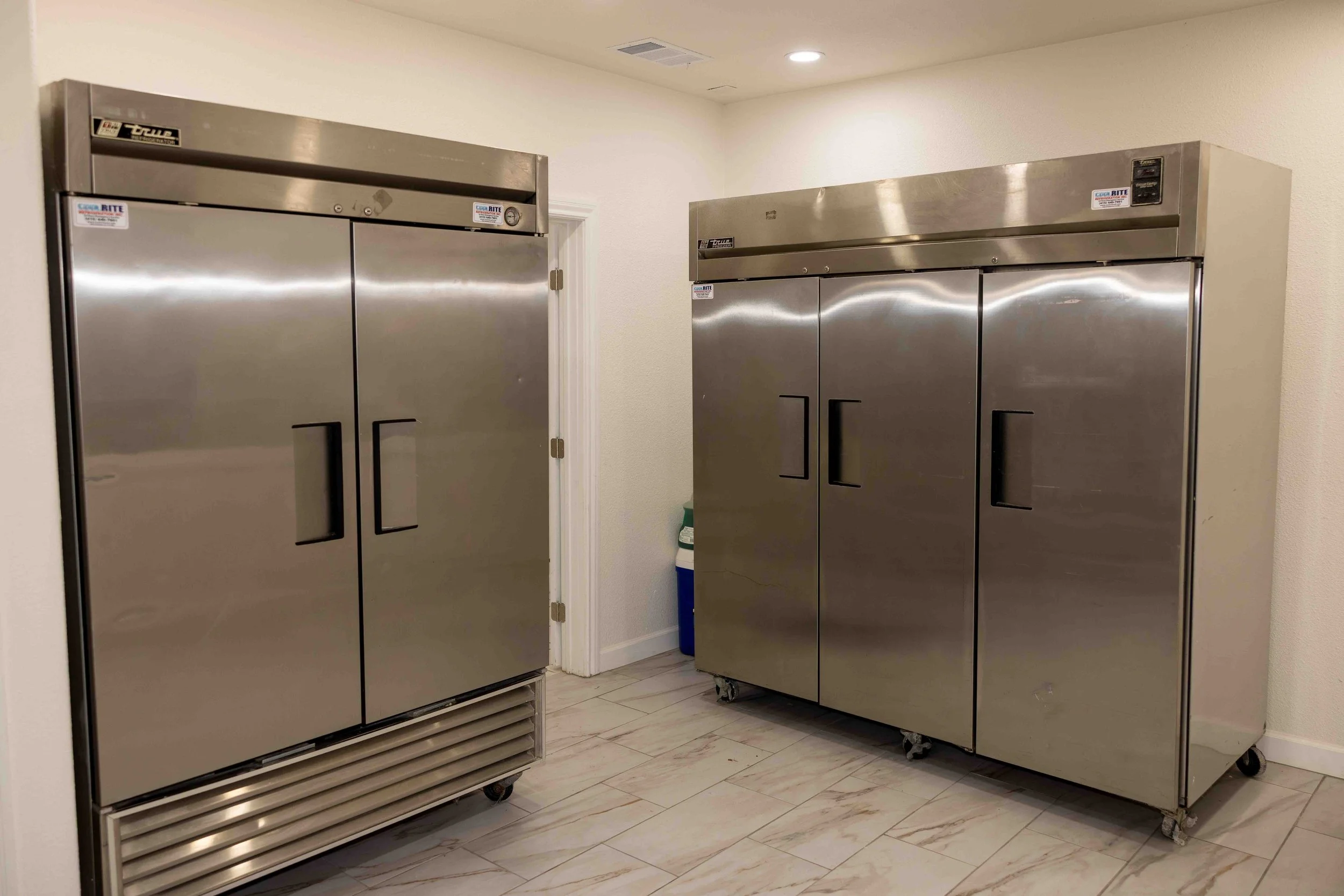 Two large stainless steel commercial refrigerators with double doors in a room with beige tiled flooring and white walls.
