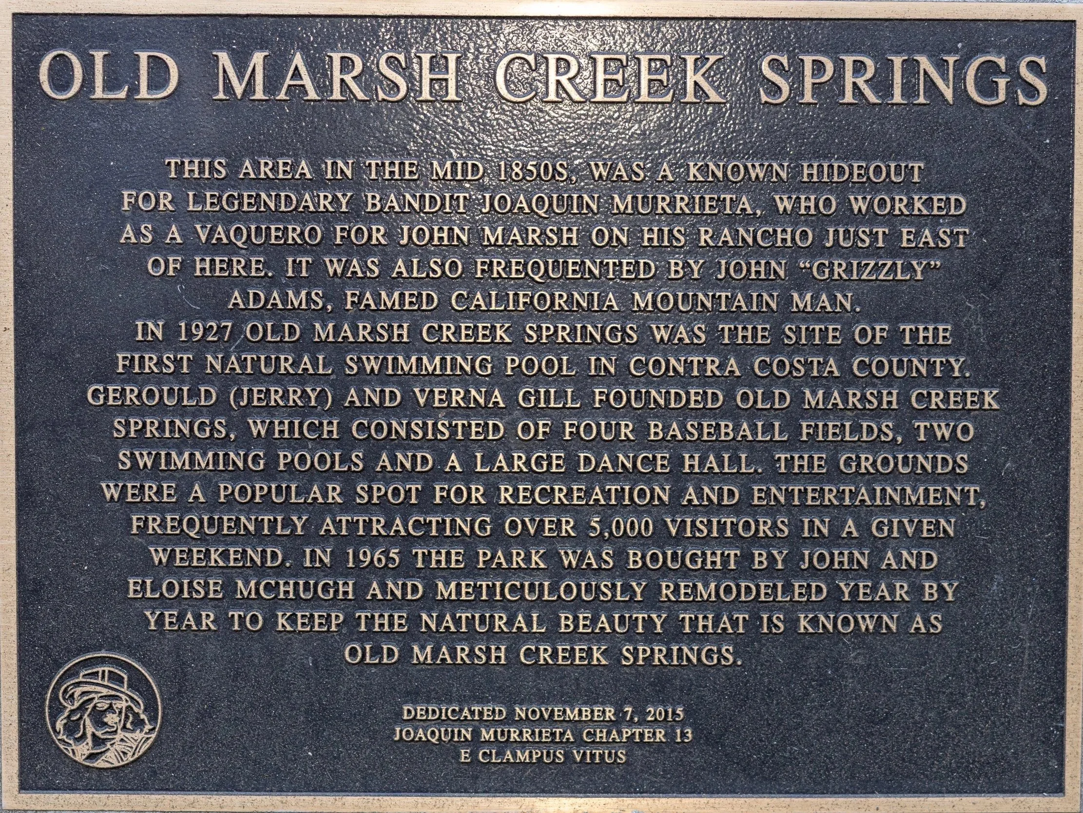 A black informational plaque with gold text about Old Marsh Creek Springs, mentioning its history from the 1850s and 1927, notable figures, and renovations in 1965, with a small logo of a person's face wearing a hat at the bottom left.
