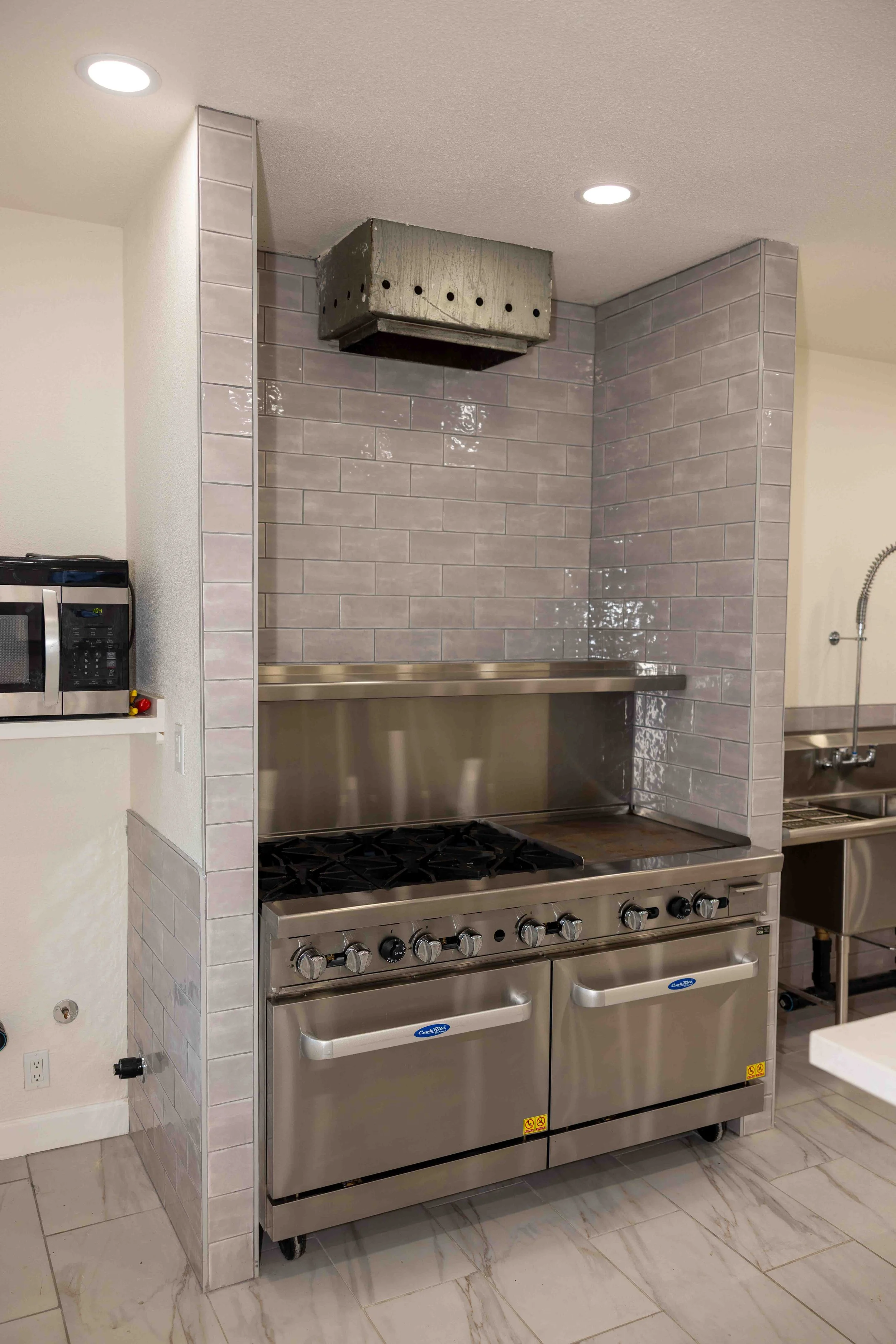 Commercial kitchen with stove, microwave, and stainless steel appliances, gray tiled wall, and tiled floor.