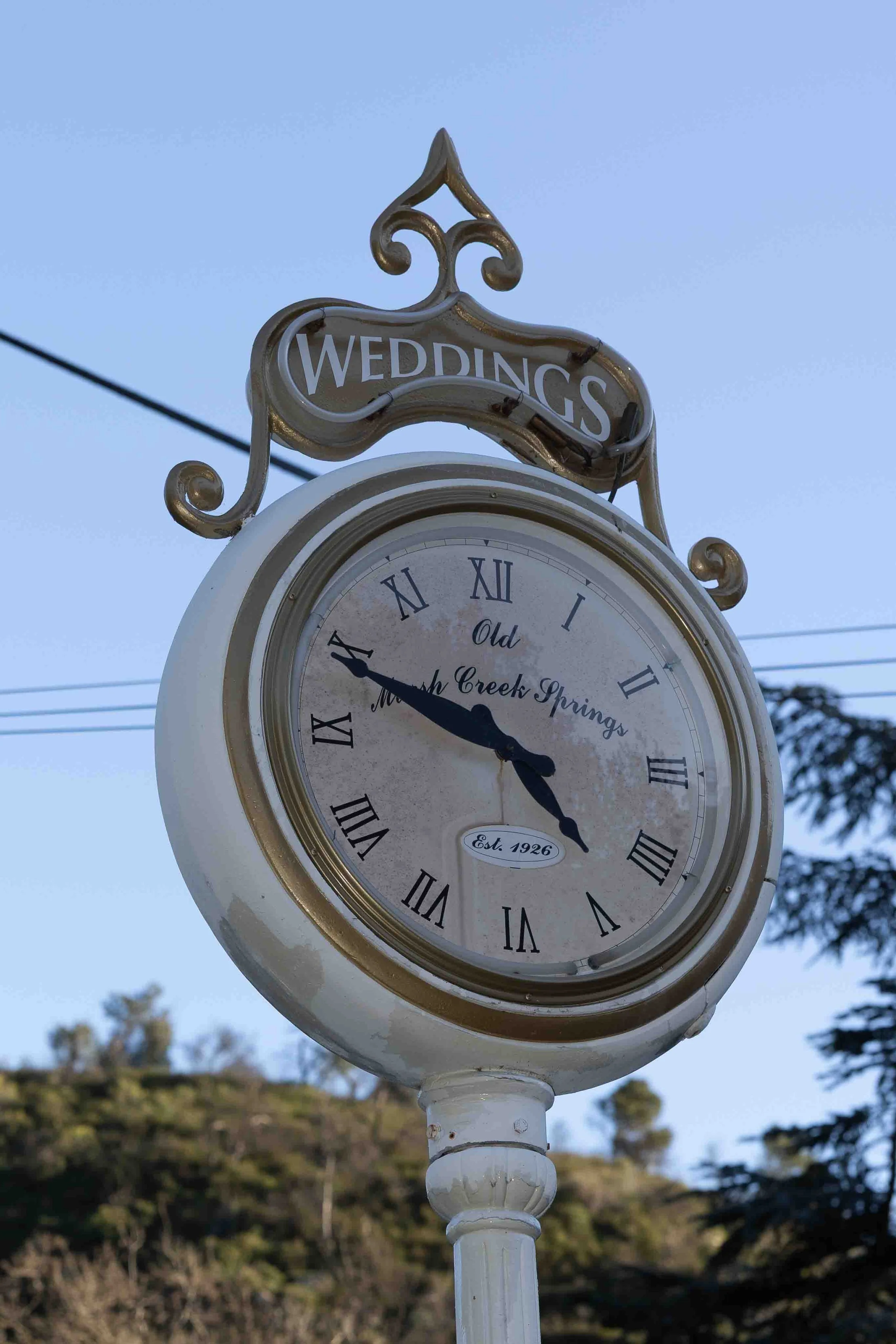 A vintage clock on a white post showing the time as approximately 4:47, with a sign above it that reads 'WEDDINGS'. The clock face has Roman numerals and the words 'Old Moon Creek Springs' and 'Est. 1926' written on it. There are power lines and trees in the background under a clear sky.