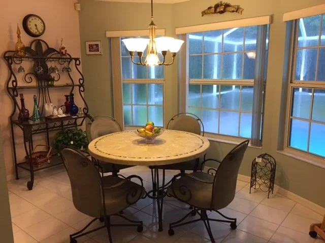 A dining room with a round table and four chairs, hanging chandelier, large windows with blinds, a decorative shelf with glass vases and a clock, and a bowl of fruit on the table.