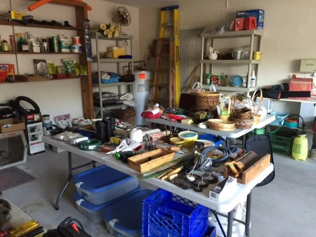 Garage or storage room with shelves filled with various household items, a table cluttered with tools, baskets, and miscellaneous objects, and storage containers on the floor.
