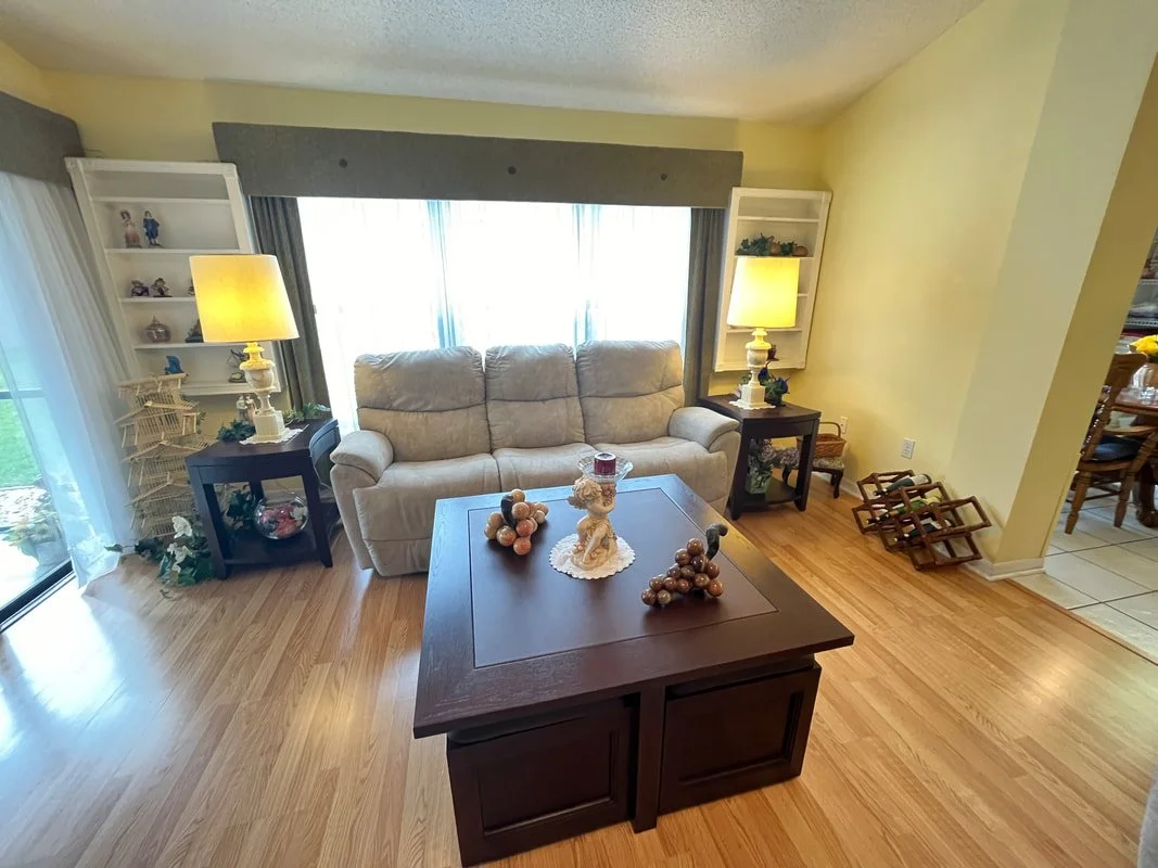 Living room with cream sofa, wooden coffee table with holiday decorations, side tables with lamps, bookshelves with decorative items, large window with curtains, hardwood flooring.