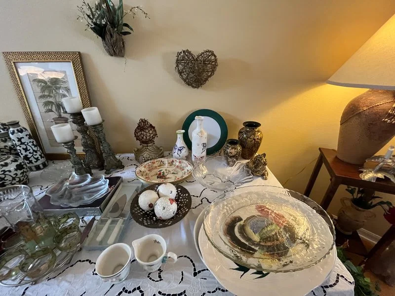 A dining table decorated with various dishes, vases, candles, and decorative items, set against a wall with heart-shaped wall art and a framed picture, with a side table and lamp nearby.