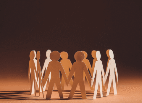 Paper cutout figures holding hands in a circle, standing on a brown surface with a dark background.