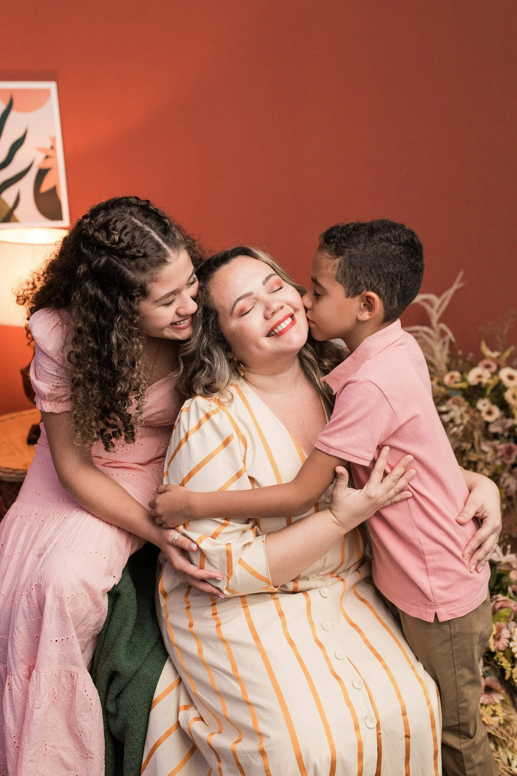 A woman with curly hair, wearing a striped dress, surrounded by two children, a girl on her left and a boy on her right, all smiling and hugging each other in a room with a warm ambiance and floral arrangements in the background.