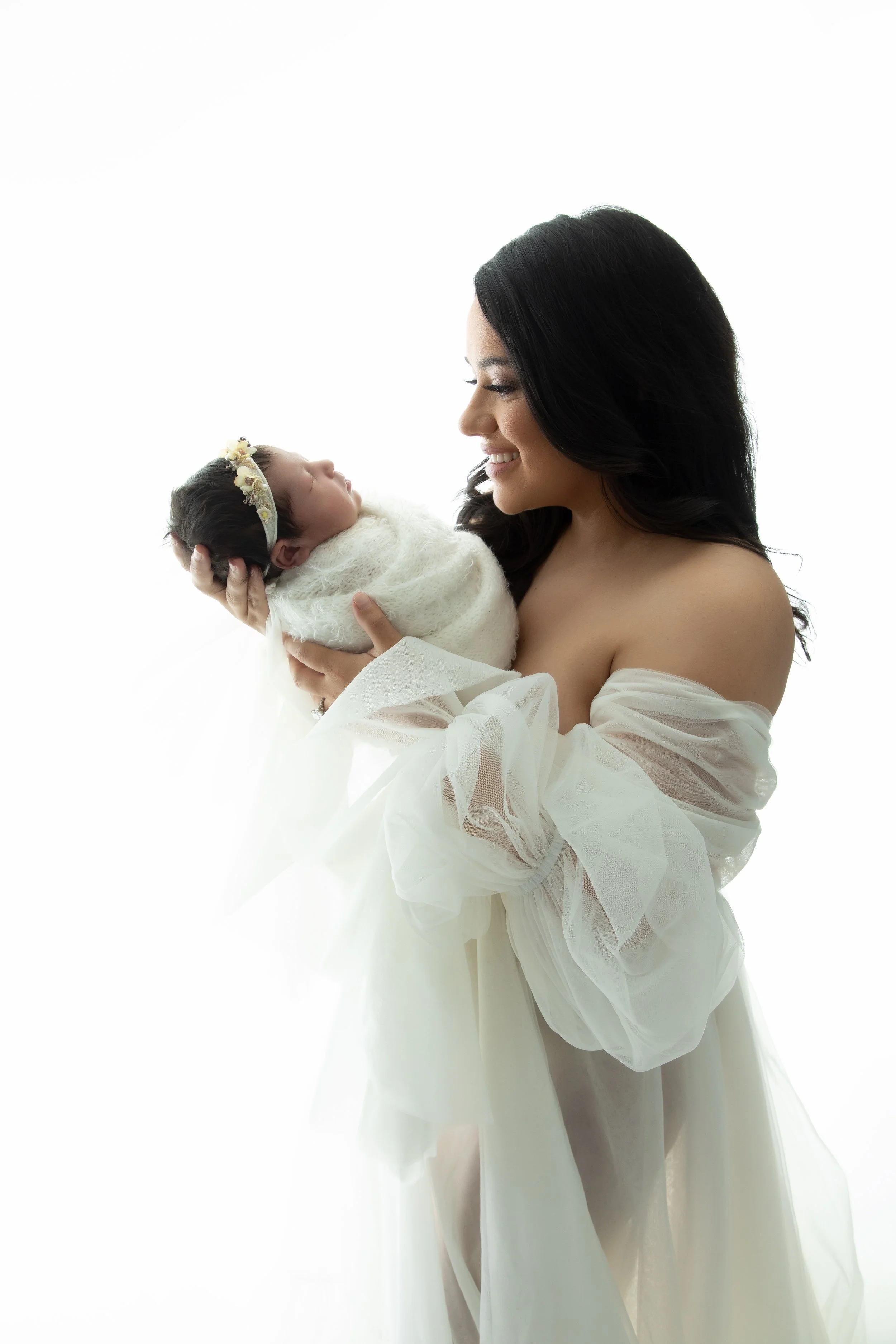 A woman with black hair holding a newborn baby wrapped in white blanket, both against a white background.