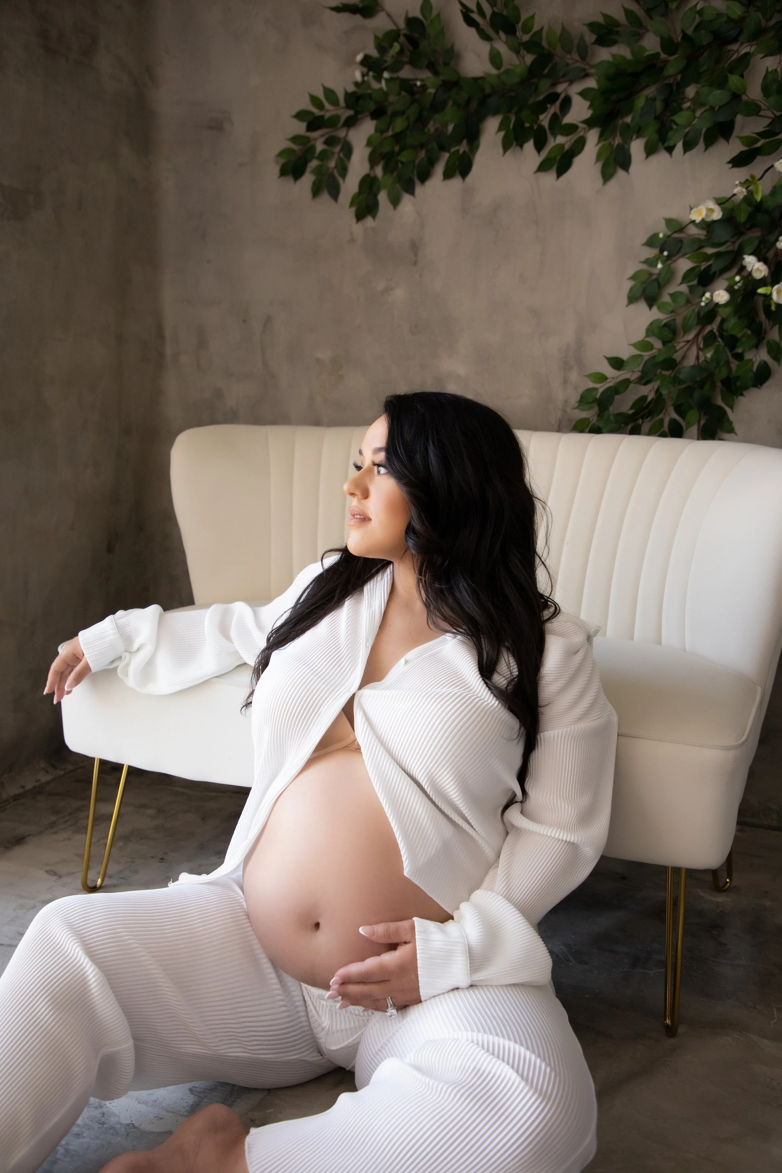 A pregnant woman with long dark hair, wearing a white ribbed outfit, sitting on the floor in front of a cream-colored tufted sofa, with a green leafy plant on a concrete wall behind her.