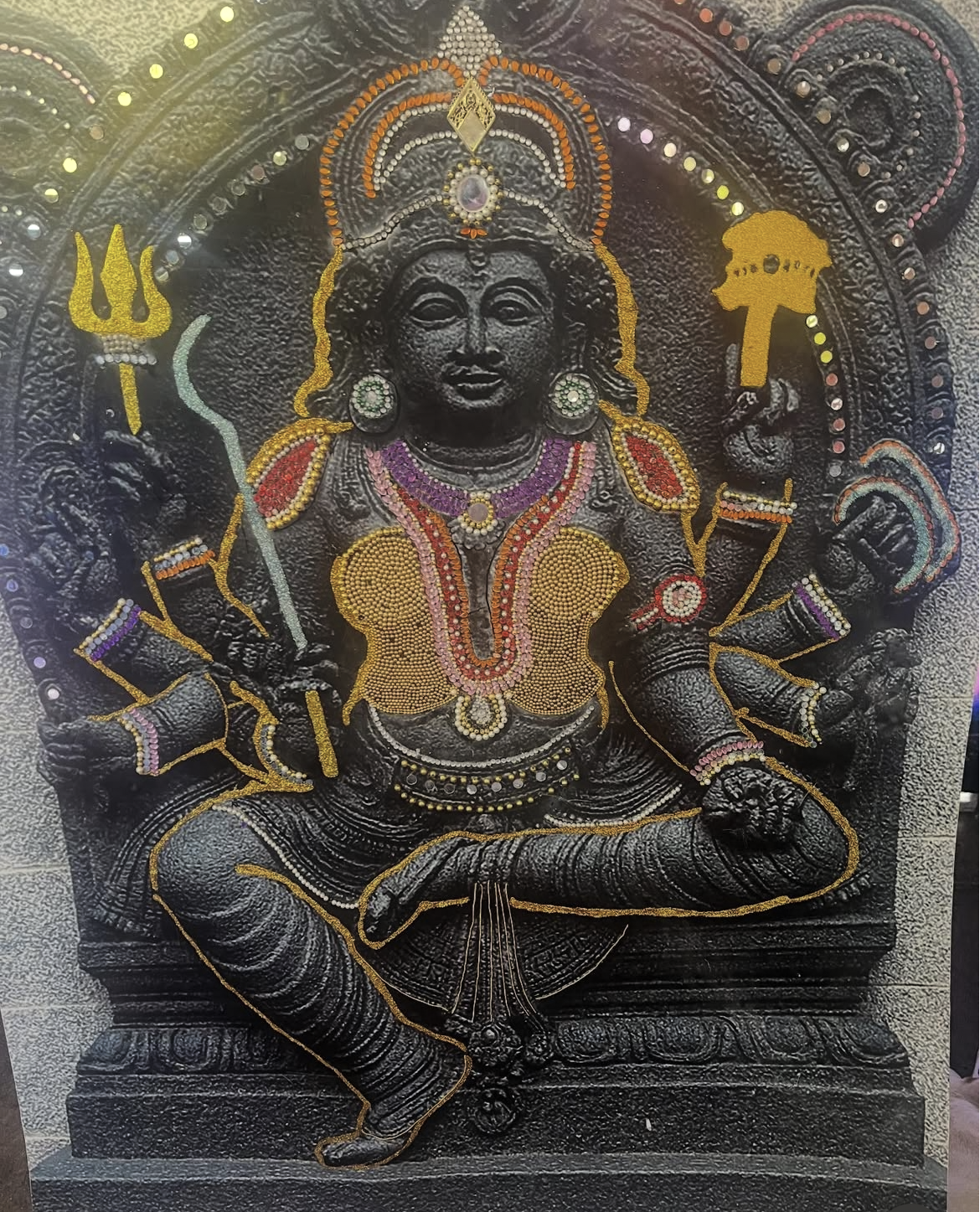 Black stone sculpture of Lord Vishnu adorned with colorful jewelry and decorations, holding a trident and a conch, with a halo of dots around the head.
