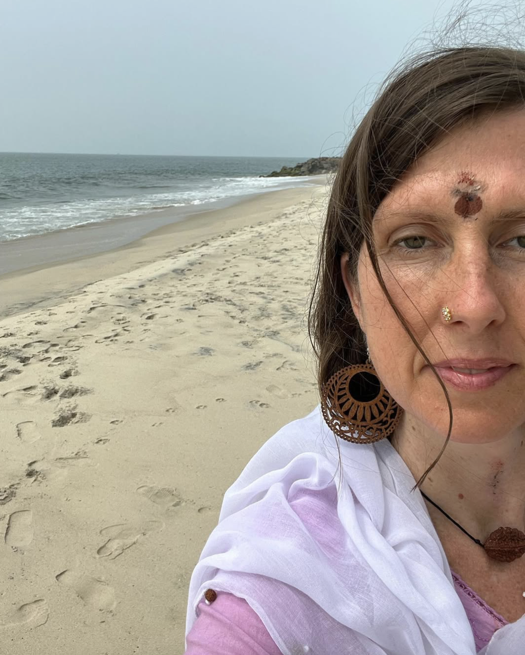 A woman with brown hair, piercings, and large earrings taking a selfie on a sandy beach with footprints, ocean waves, and cloudy sky in the background.