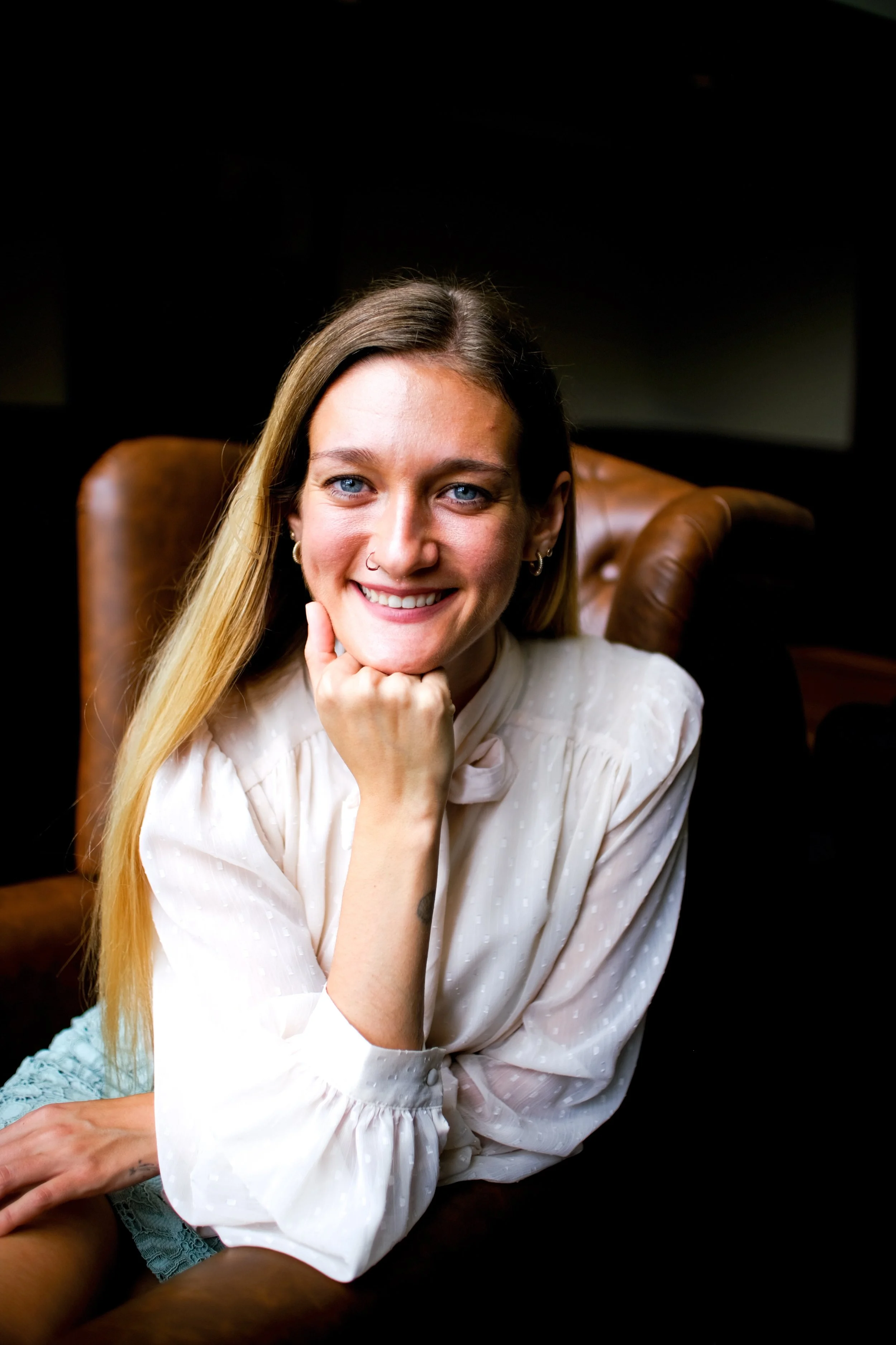 A woman with long blonde hair, blue eyes, and a nose piercing smiling while sitting on a brown leather chair, wearing a white long-sleeve blouse with subtle polka dots.