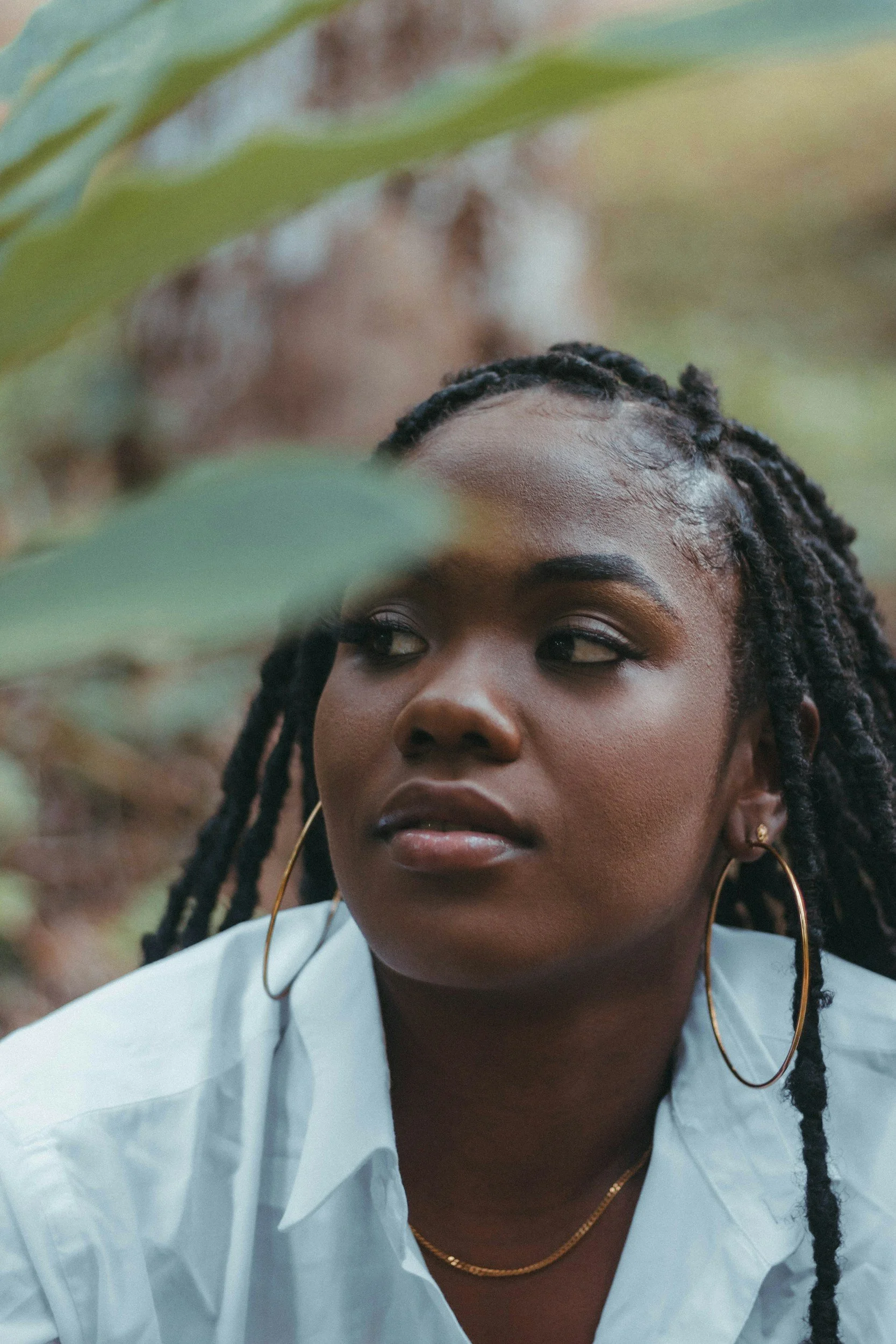 A young woman with braided hair and gold jewelry looks thoughtfully to the side amidst lush green foliage.