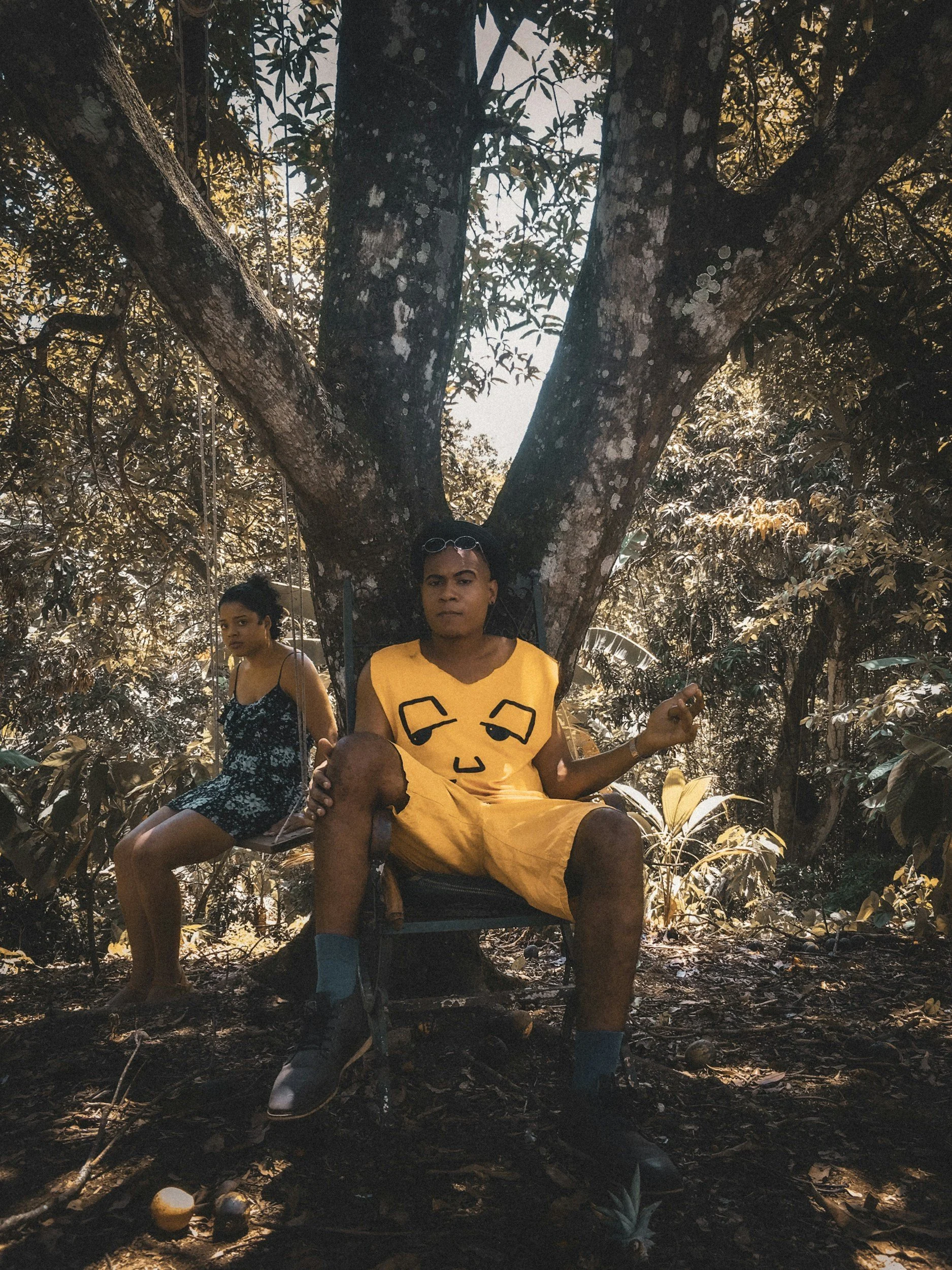 Two people sitting in a tree in a forest, one person in yellow clothing in front, and another in a black dress behind.