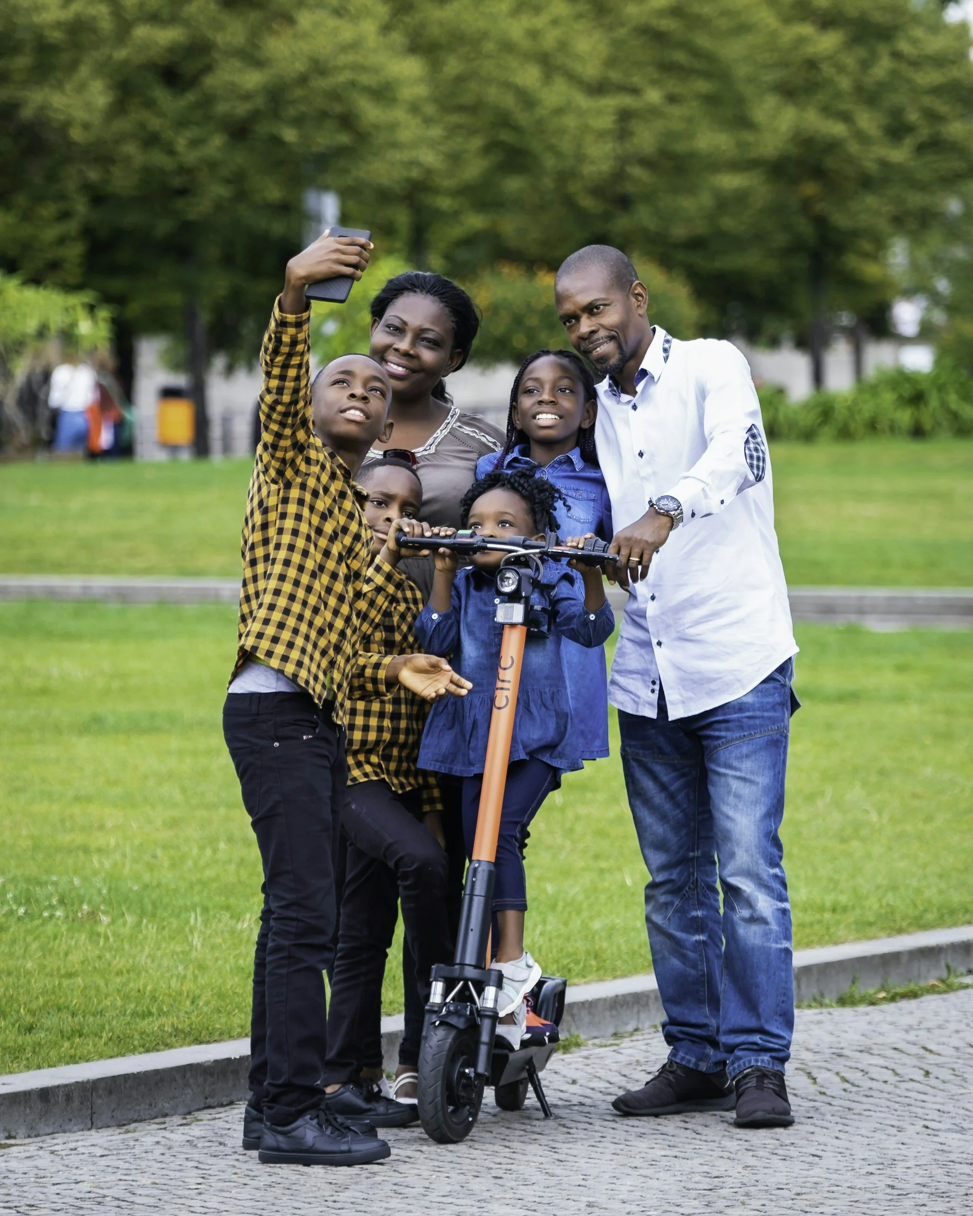 A family of five taking a selfie in a park. The family includes a man, a woman, and three children, one of whom is on a scooter. They are smiling and standing on a paved path with grass and trees in the background.