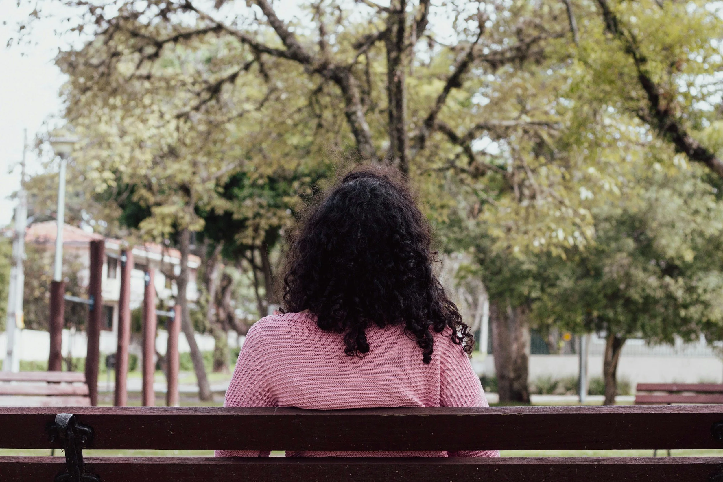 A woman with curly black hair, wearing a pink sweater, sitting on a park bench facing away, with trees and a lamppost in the background.