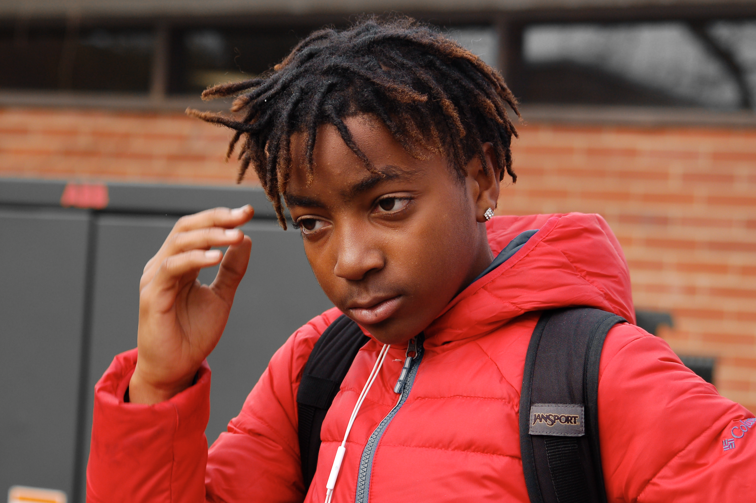 Young man with dreadlocks wearing a red jacket, black backpack, and earbuds, standing outdoors in front of a brick building and gray lockers, adjusting his hair or forehead.