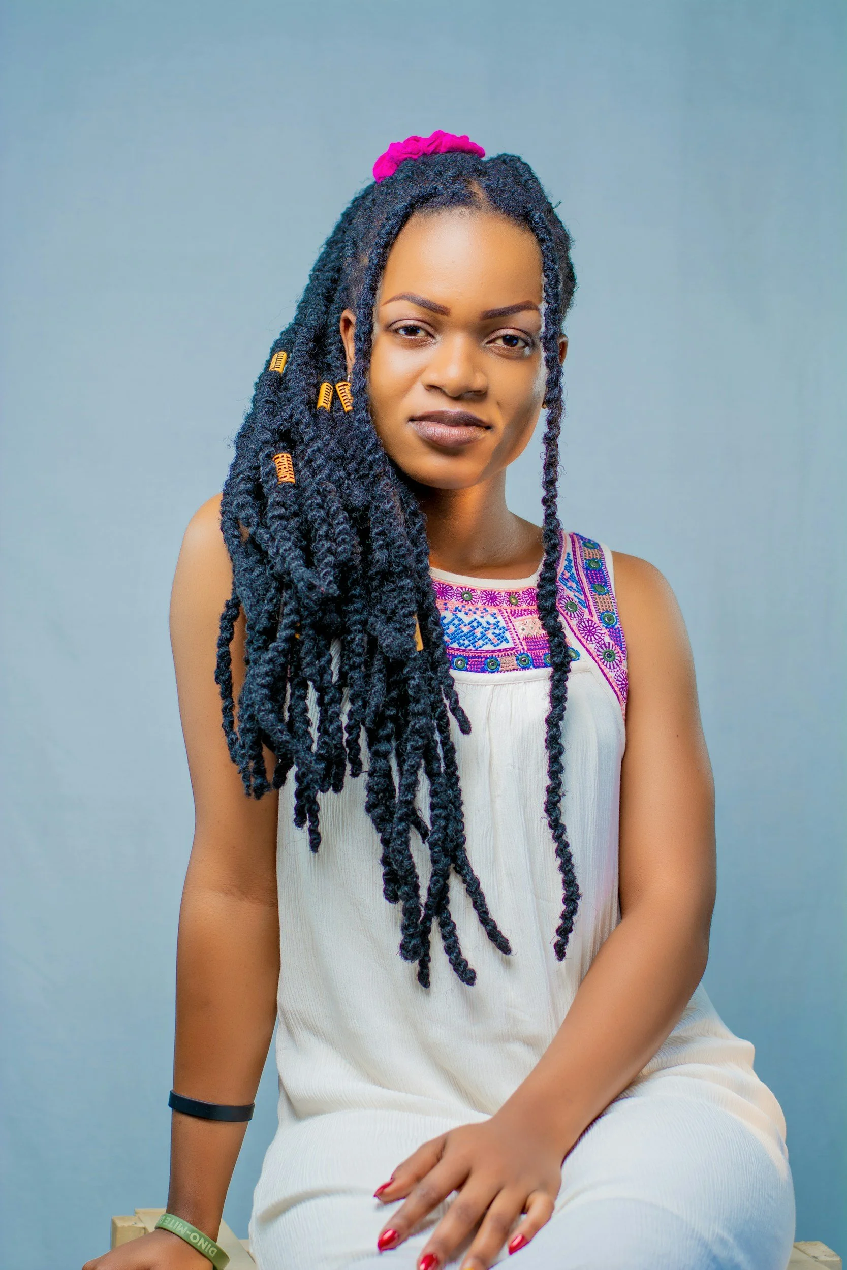 A woman with styled dark blue, curled box braids adorned with colorful beads, wearing a white dress with colorful embroidered details around the neckline, sitting against a light blue background.