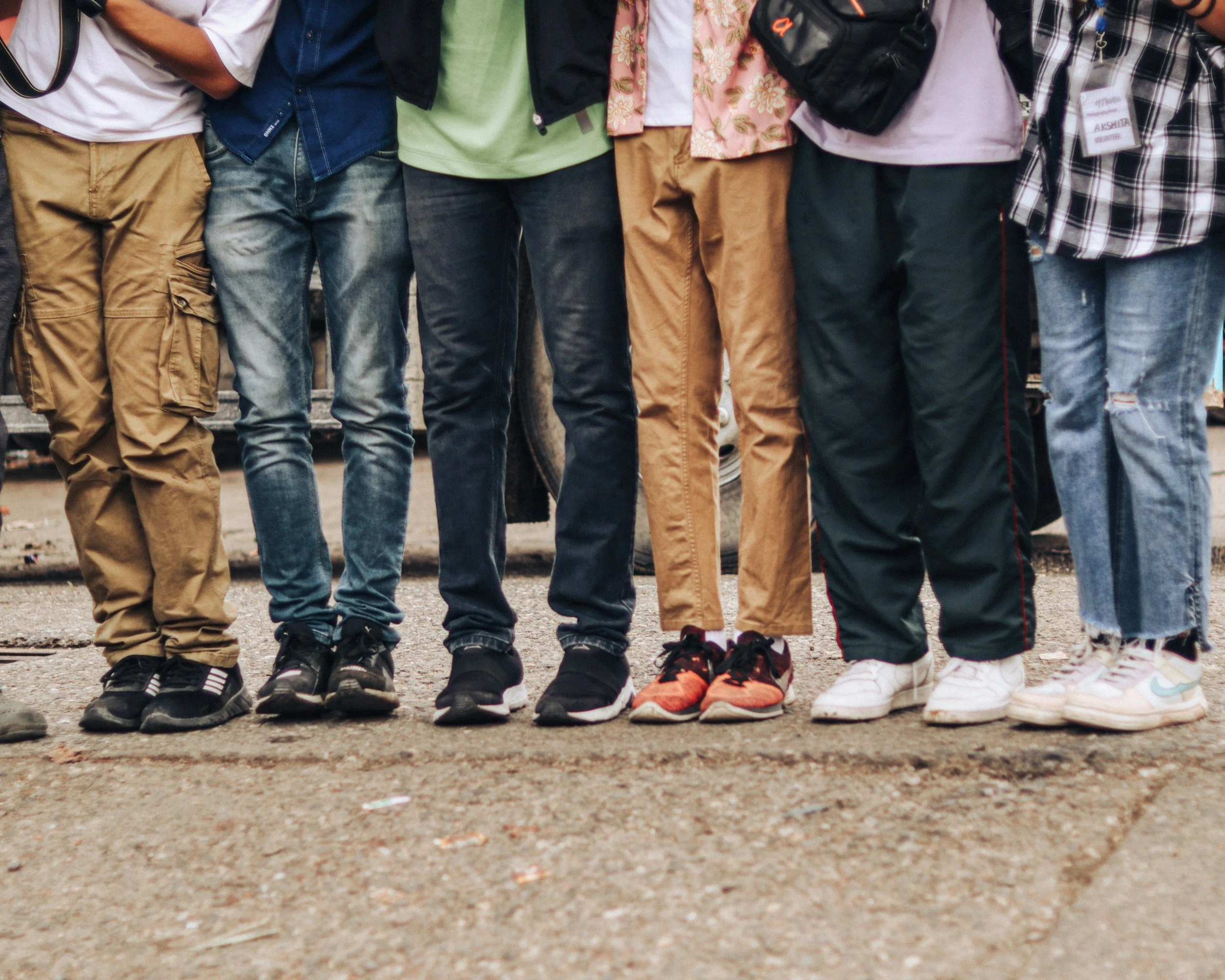 The image shows the lower halves of a group of people standing closely together outdoors, wearing casual clothing and sneakers.