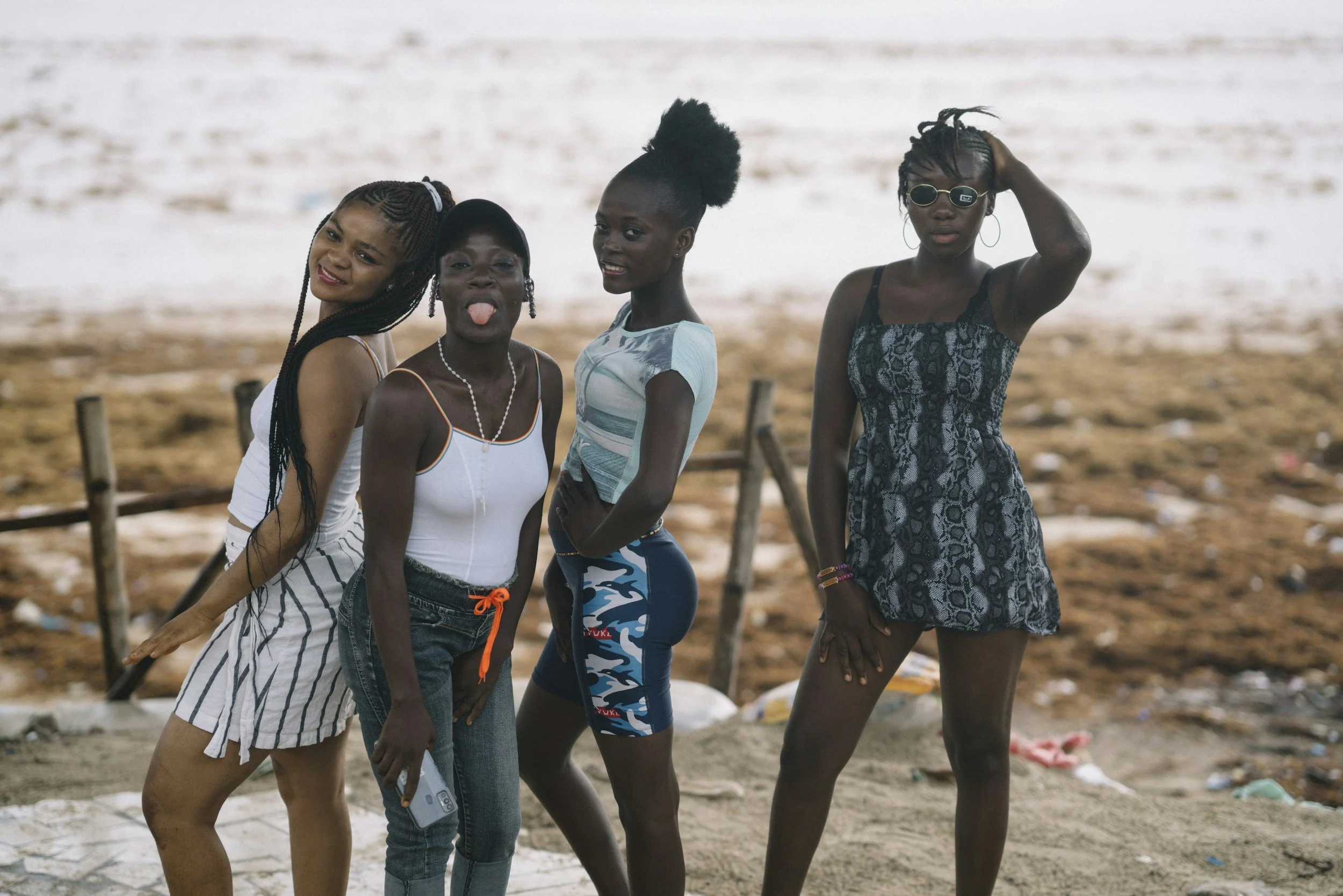Four young women are standing outdoors on a sandy area with trash and wooden posts in the background. They are posing for the photo, making playful expressions and gestures. The first woman on the left is wearing a striped skirt and tank top, smiling and leaning slightly. The second woman is sticking her tongue out, wearing a white top and jeans. The third woman is with her hand on her hip, wearing a blue patterned sports outfit, smiling. The fourth woman is wearing sunglasses and a black and gray dress, touching her hair.
