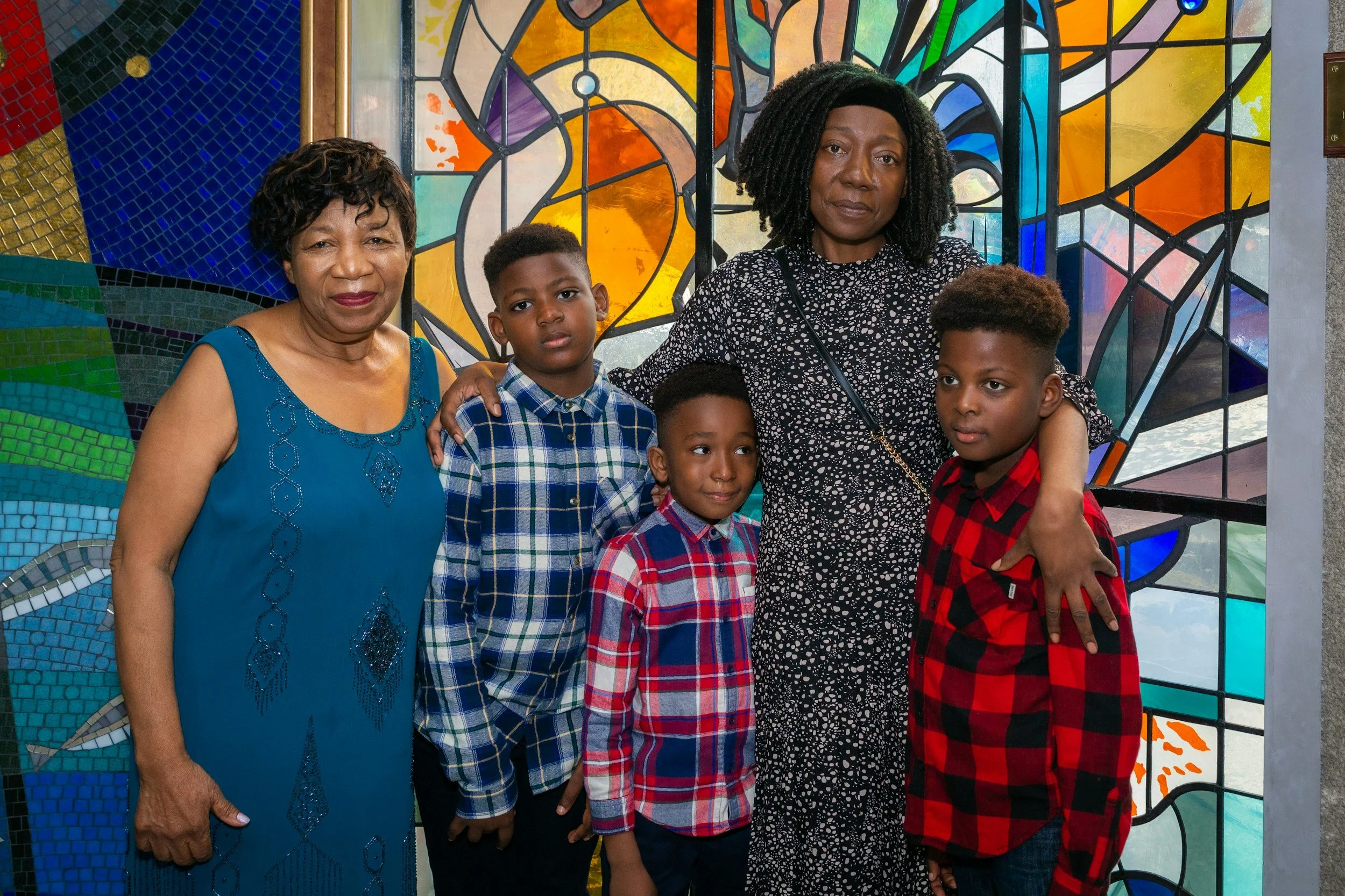 A group of five people standing in front of colorful stained glass windows, including two older women and three children, all looking at the camera.