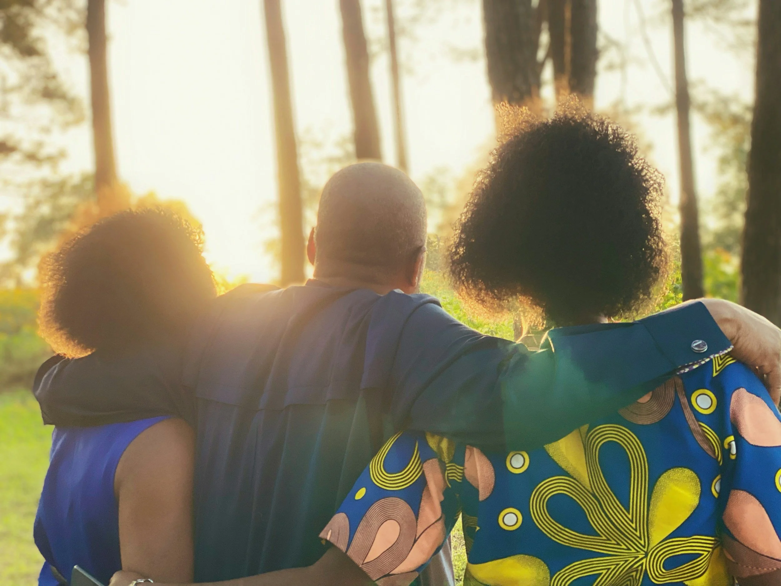 Three people, two women and one man, sitting close with arms around each other, watching a sunset in a forest.
