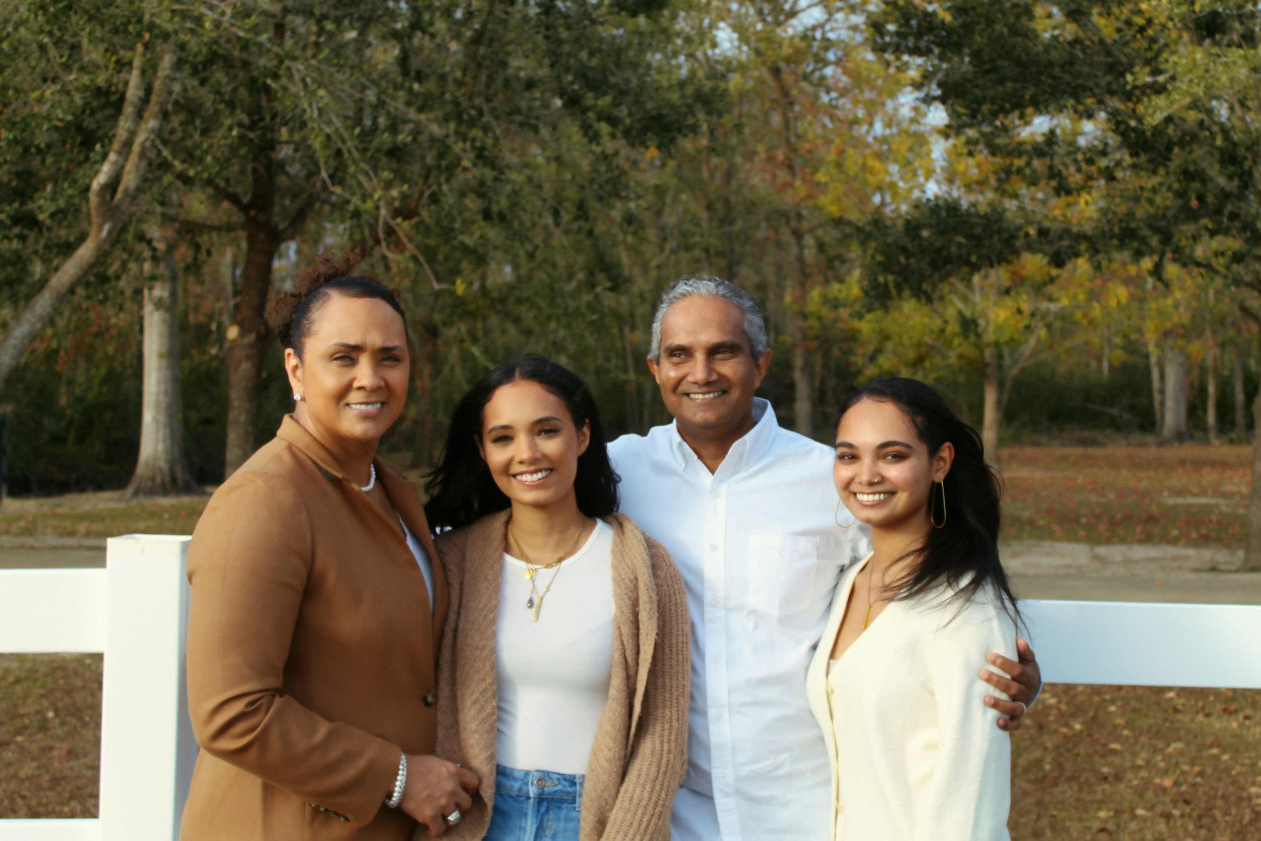 Family of four smiling outdoors in a park during fall, standing in front of a white fence with trees and autumn foliage in the background.