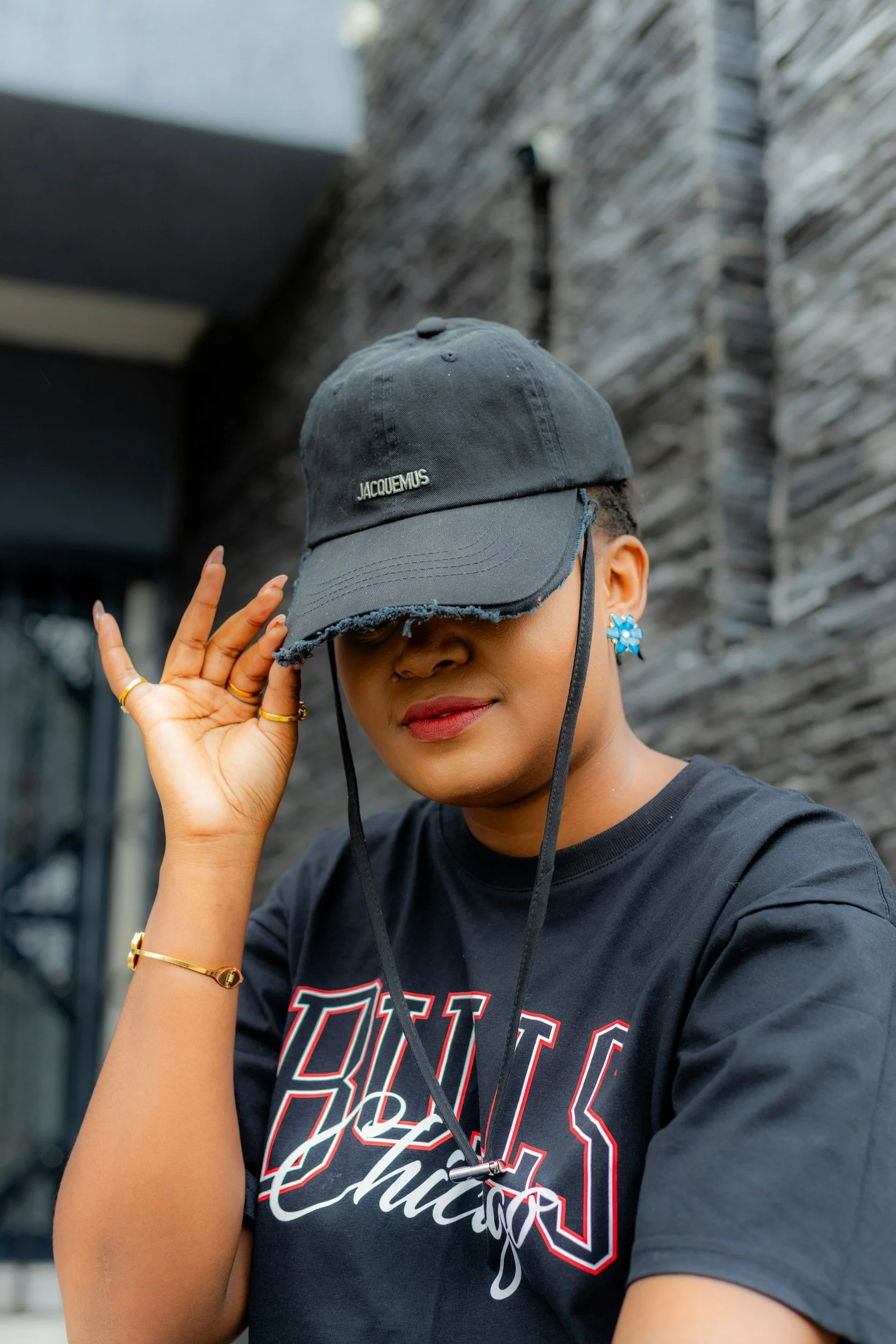 A woman wearing a black T-shirt with red and white lettering, a gray jacquemus baseball cap, and blue flower earrings, standing outdoors against a stone wall, adjusting her cap asking what is self esteem
