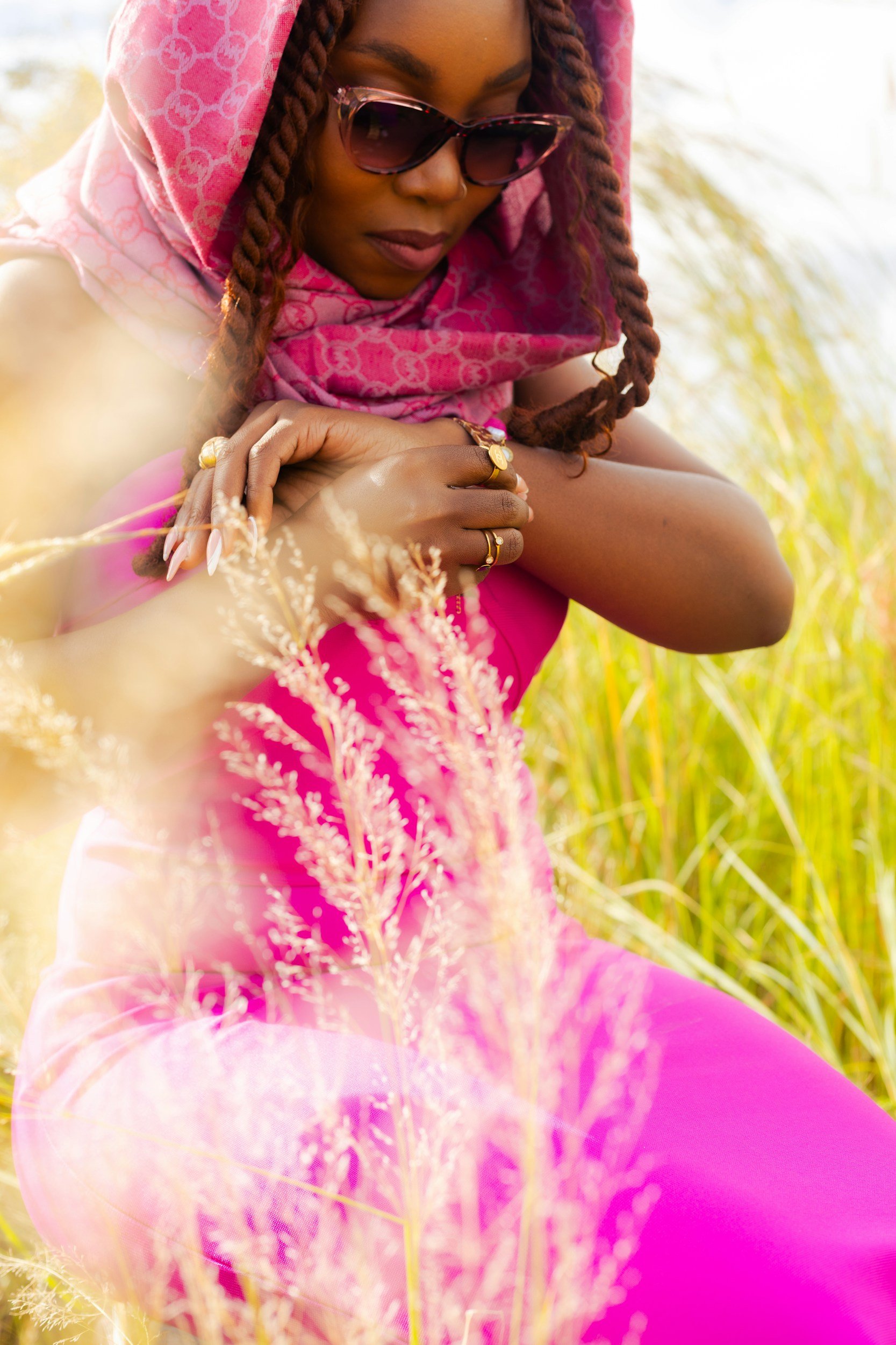 A woman wearing sunglasses, a pink scarf, and rings on her fingers, sitting in a field of tall grass, with sunlight illuminating the scene thinking about perimenopause emotional changes