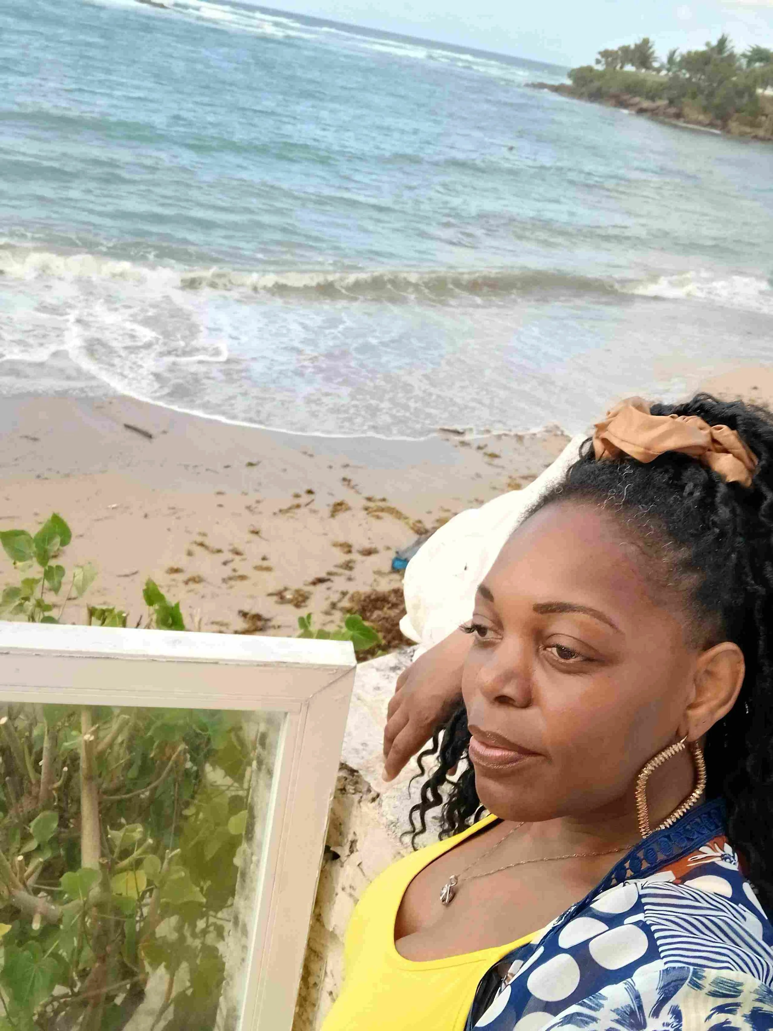 Woman with curly hair and hoop earrings looking down at a glass display case with plants near the beach with waves and sandy shoreline in the background.