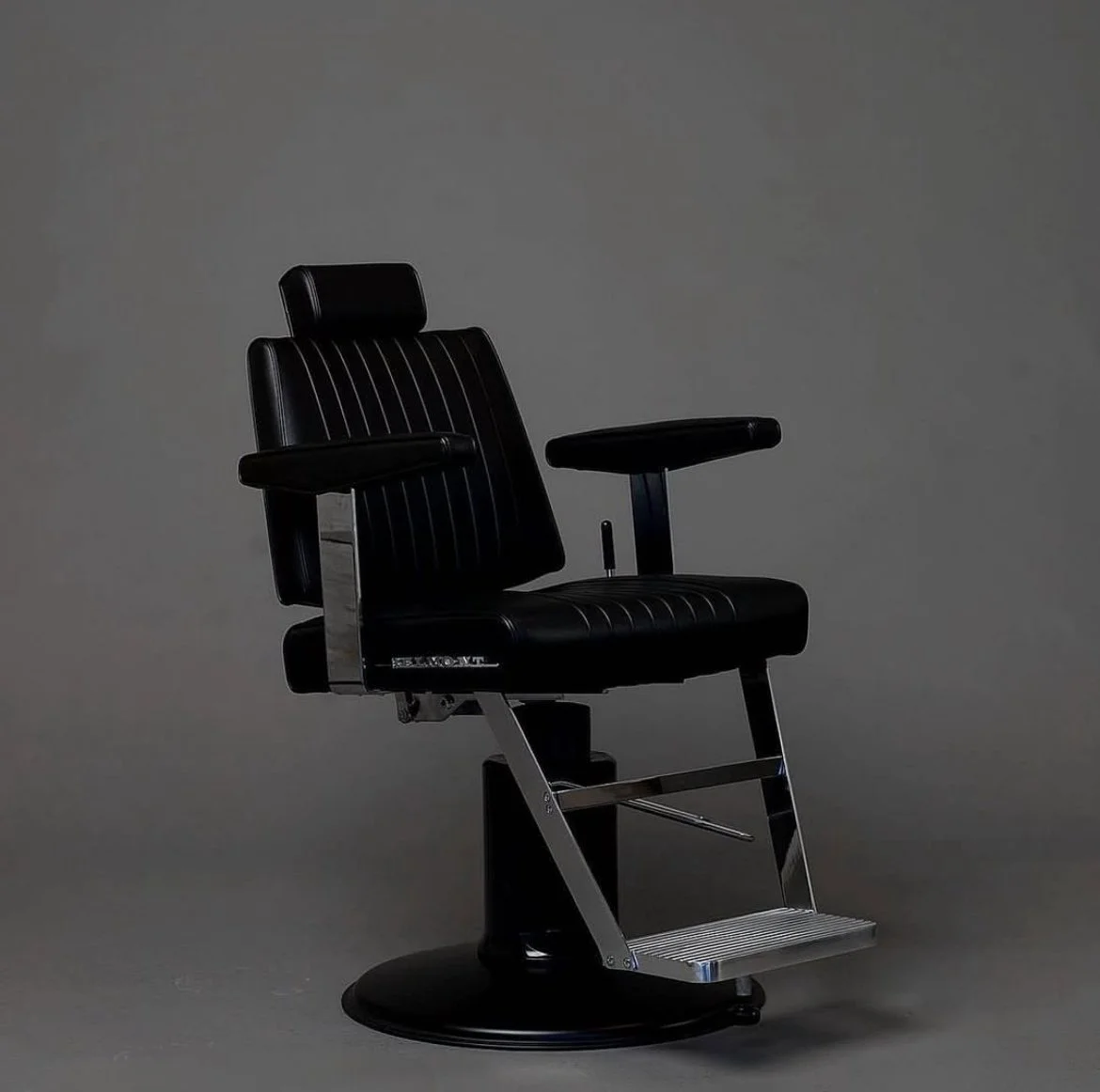 A black vintage barber chair with a rotating base, armrests, and a footrest, set against a plain gray background.