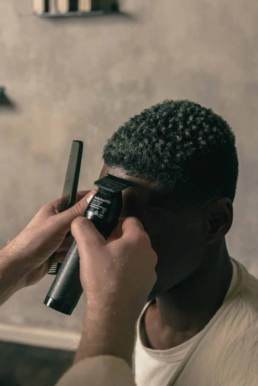A person is getting a haircut with electric clippers and a comb, focusing on the side of their head, in a barbershop or salon setting.