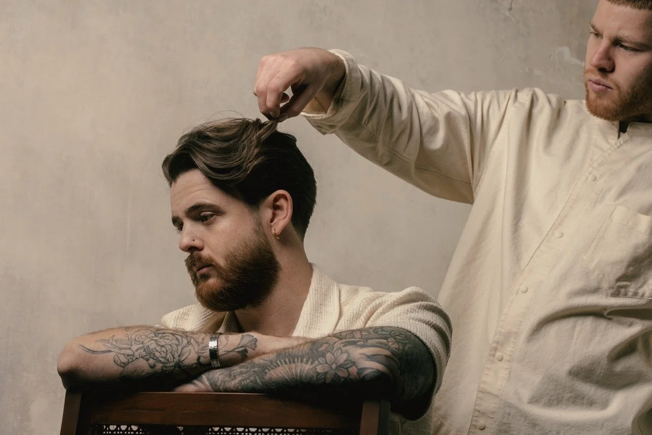 A hairstylist is cutting a man's hair in a salon. The man has tattoos on his arms and a beard, and he is sitting with his arms resting on a chair.
