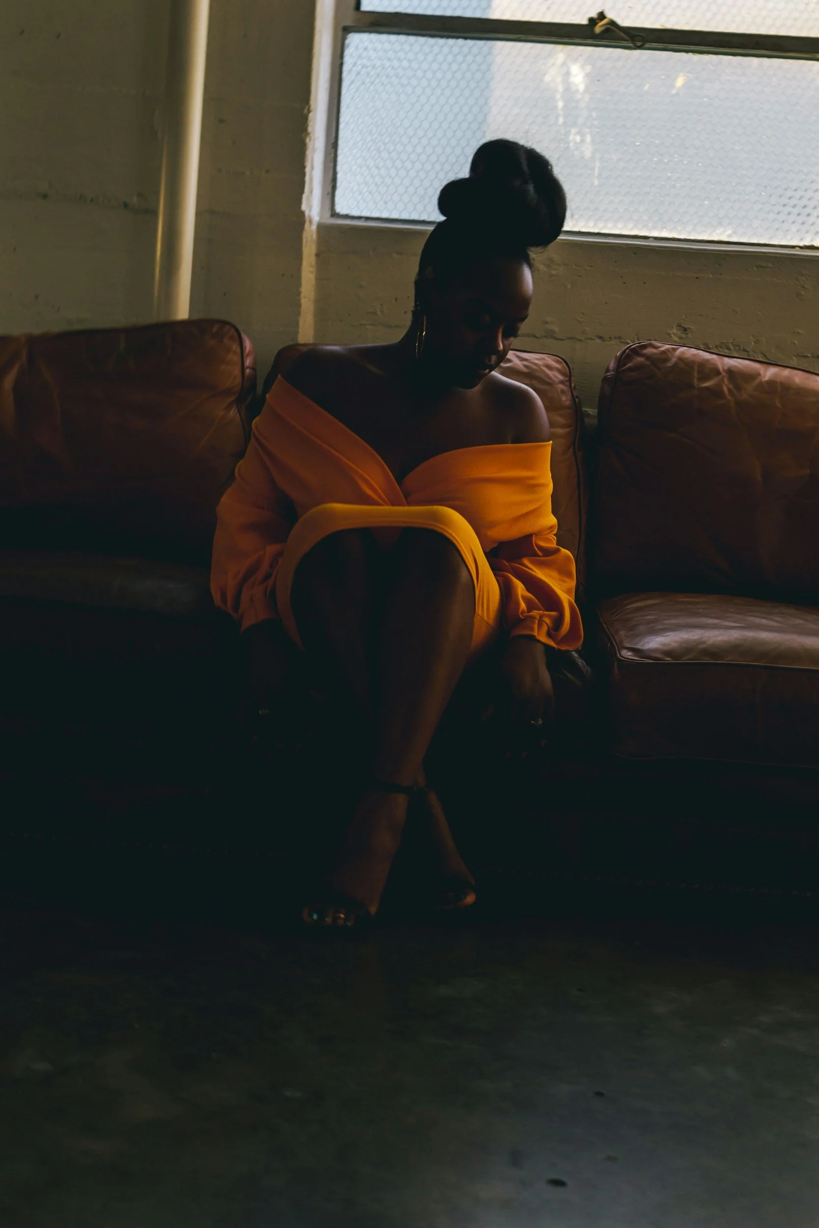 A woman sitting on a leather couch near a window with wire mesh, wearing a yellow off-the-shoulder top and high heels, with her head bowed and her hair styled in a large bun.