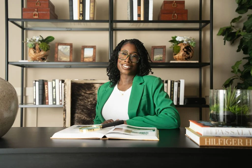 A woman with glasses and curly hair wearing a green blazer sits at a desk in an office, smiling. There are books, plants, and decorative items behind her.