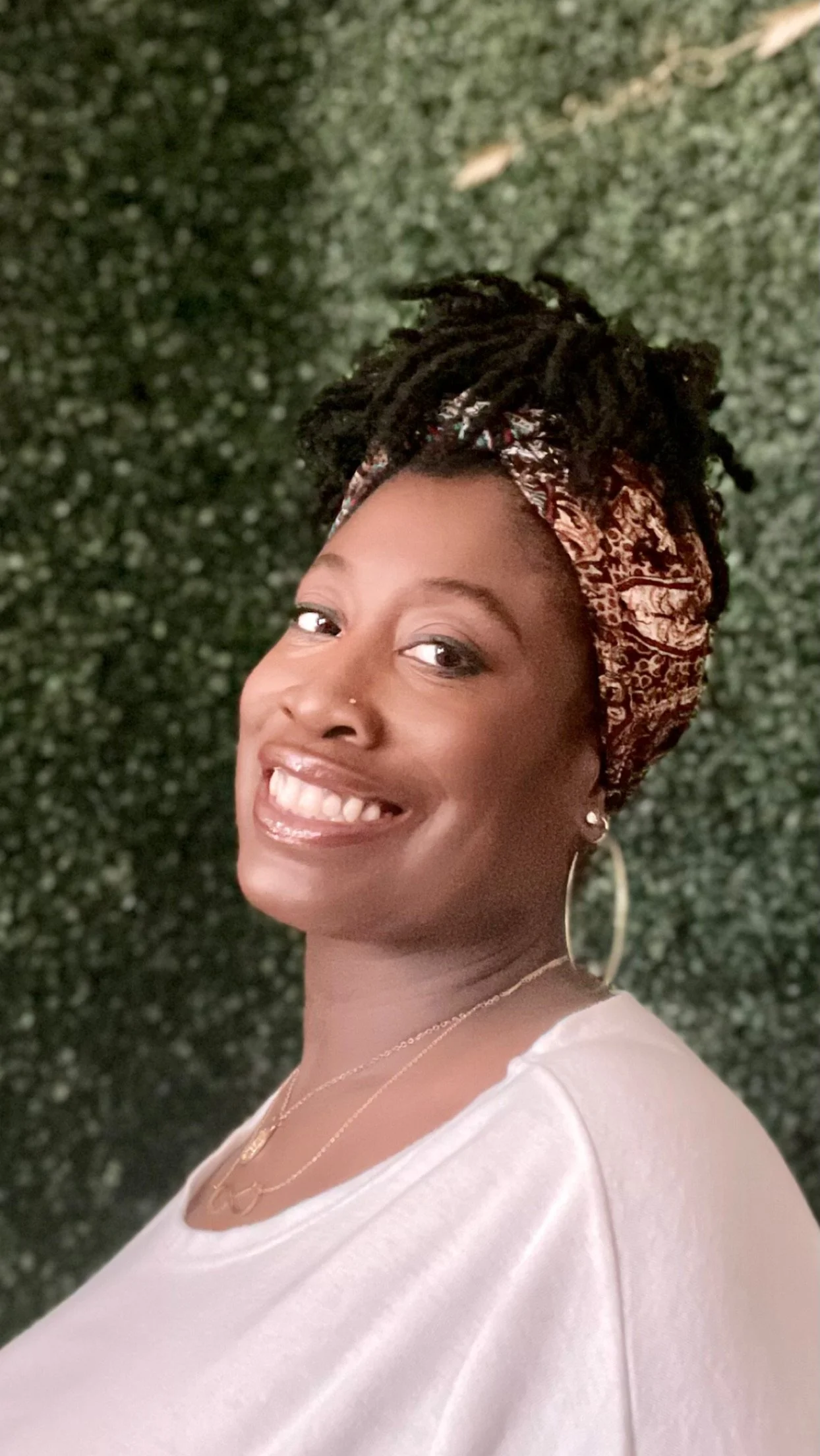 A smiling woman with dreadlocks wearing a patterned headscarf, hoop earrings, and a necklace, standing in front of a green leafy background.