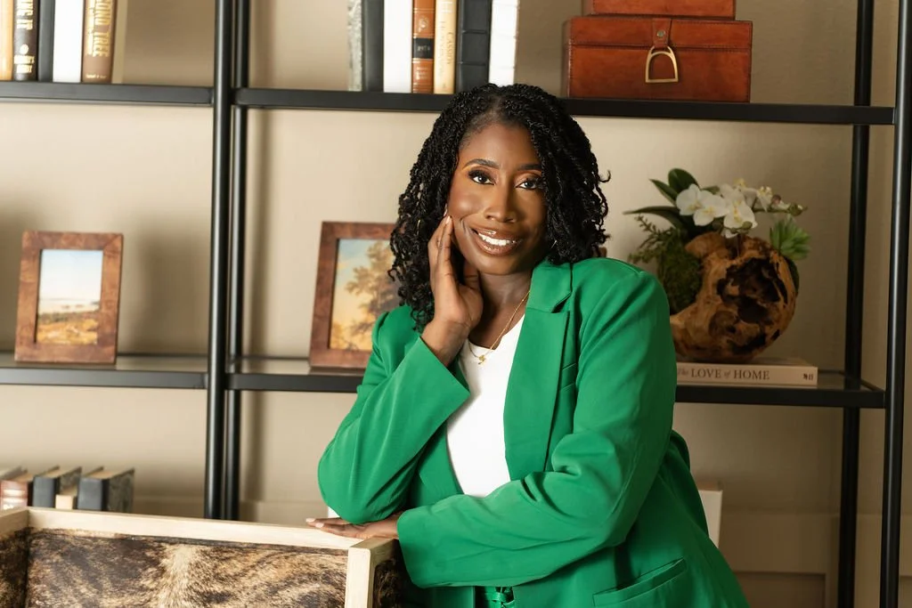A smiling woman with curly black hair wearing a green blazer and white shirt, posing indoors in front of a bookshelf with decorative items and framed photos.