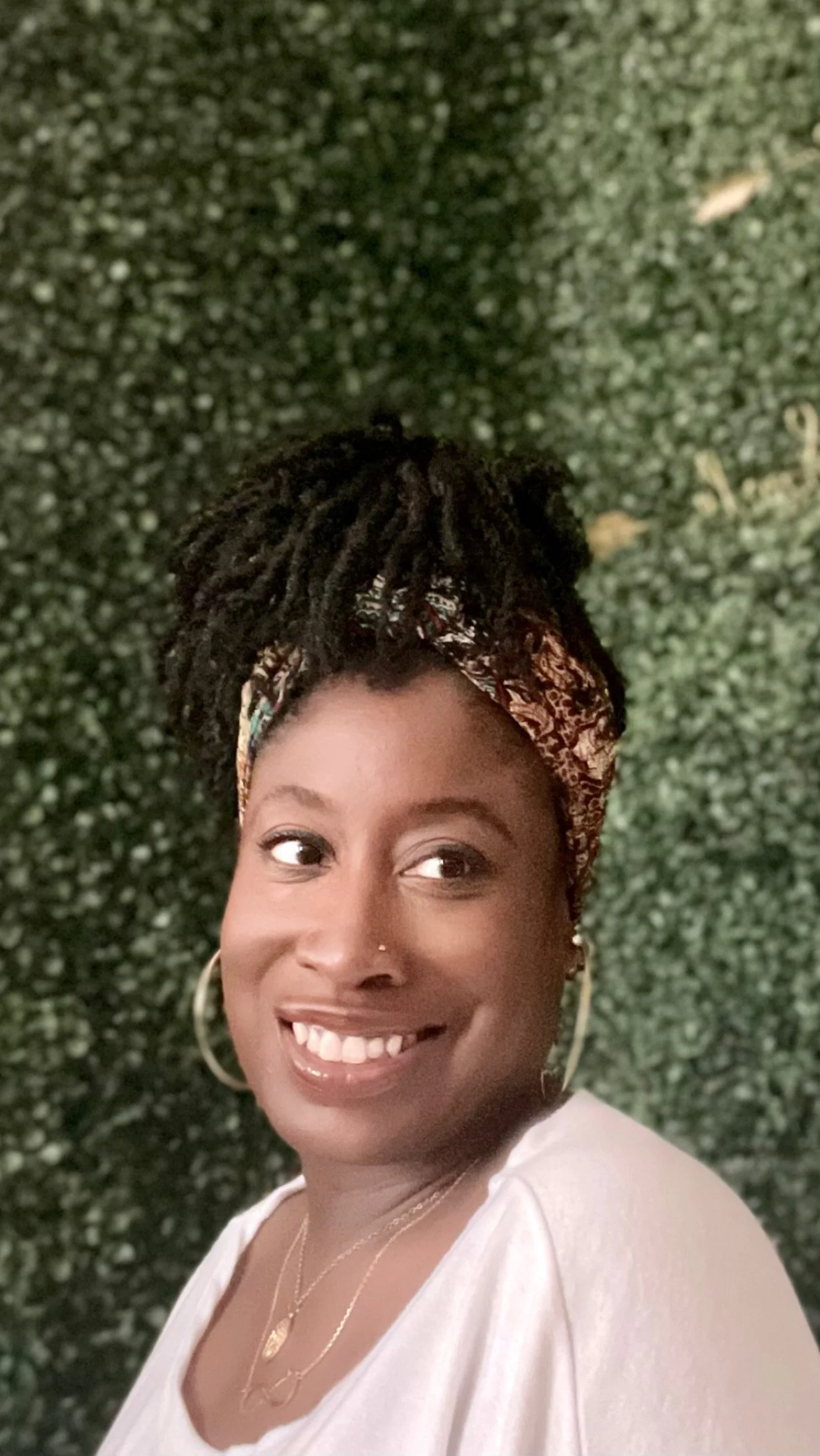 A smiling woman with dark skin, wearing a floral headscarf, hoop earrings, and layered necklaces, standing in front of a green leafy background.