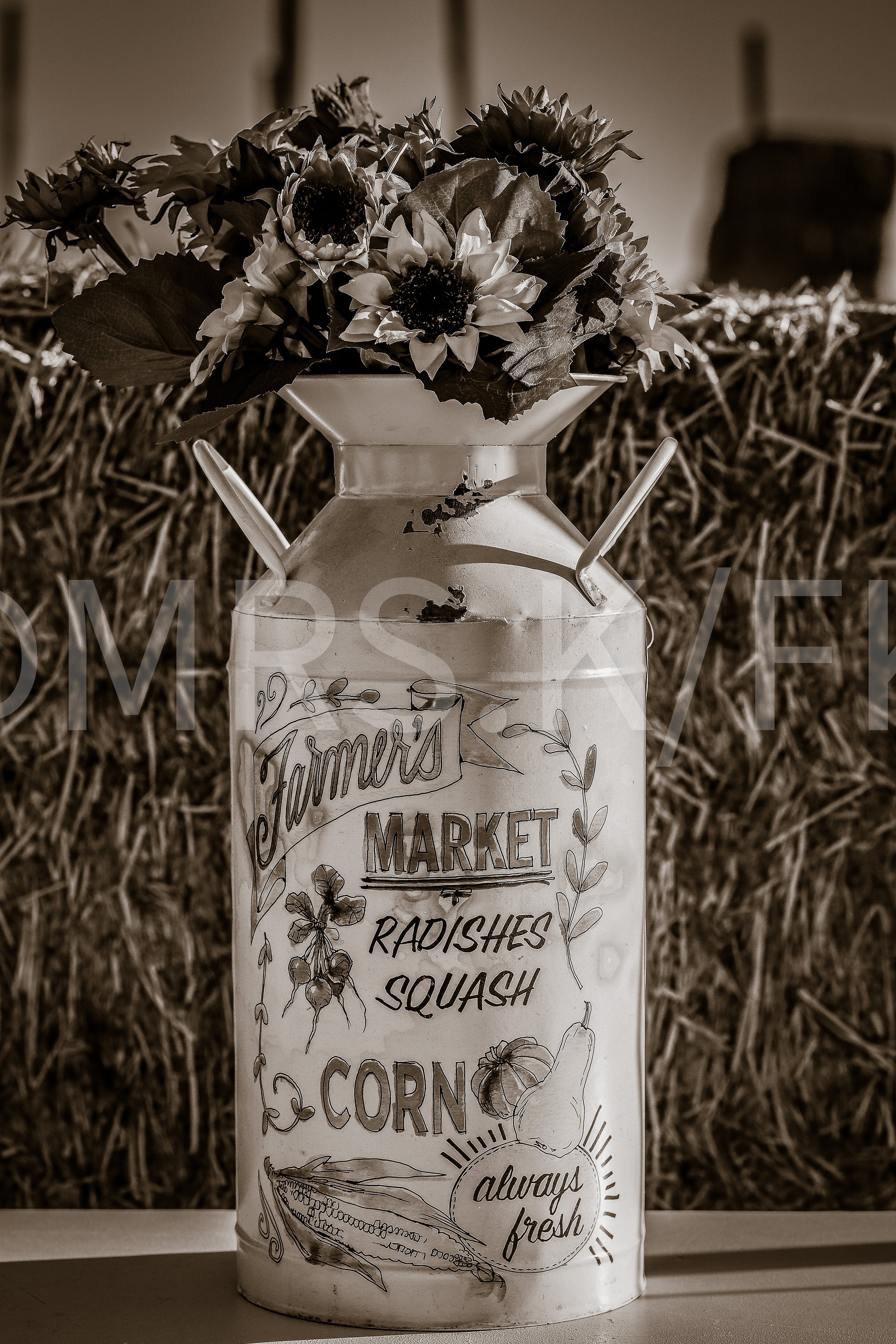 A vintage milk can with hand-drawn illustrations and text advertising farm-fresh market vegetables like radishes, squash, and corn, placed on a table against a background of hay or straw.