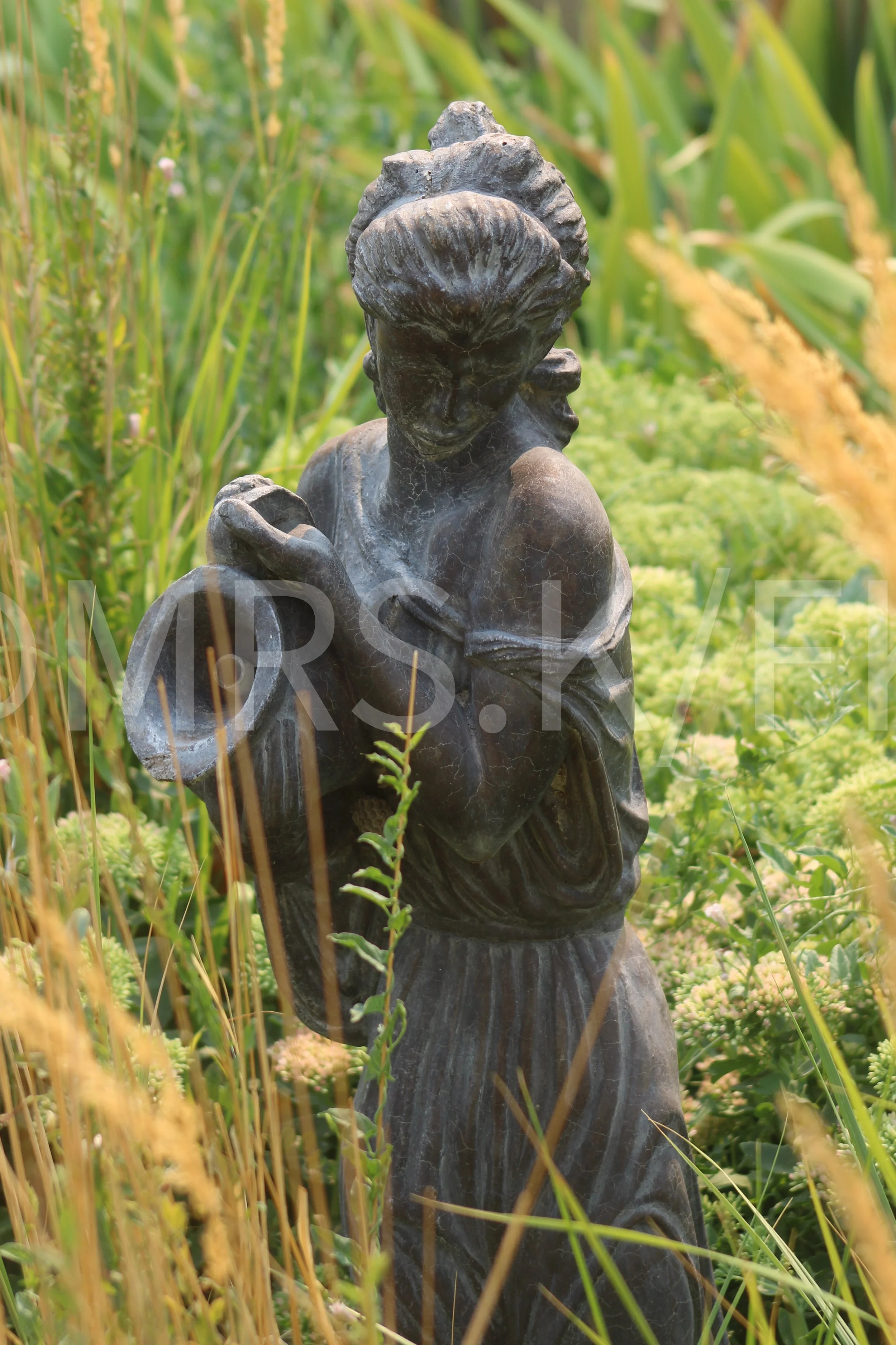 An Original Piece of Photography 1 of 10 to print Custom Fine Art - A dark stone sculpture of a woman with her hair styled in an updo, holding a bowl or vessel, surrounded by green plants and tall grass.