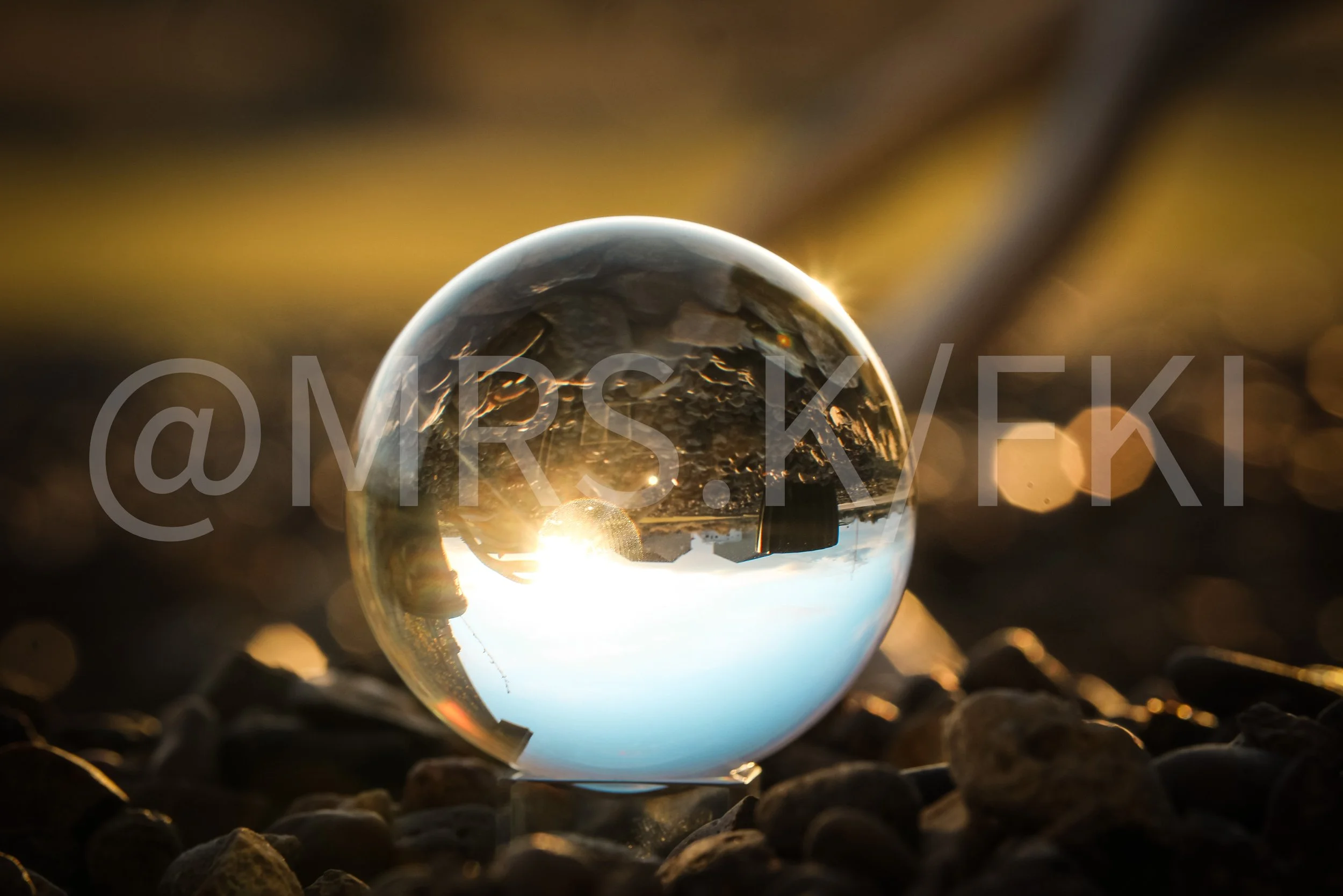 Professional Image with Water Marks, displaying a Close-up of a glass sphere reflecting a sunset scene, with rocks on the ground and bokeh lights in the background