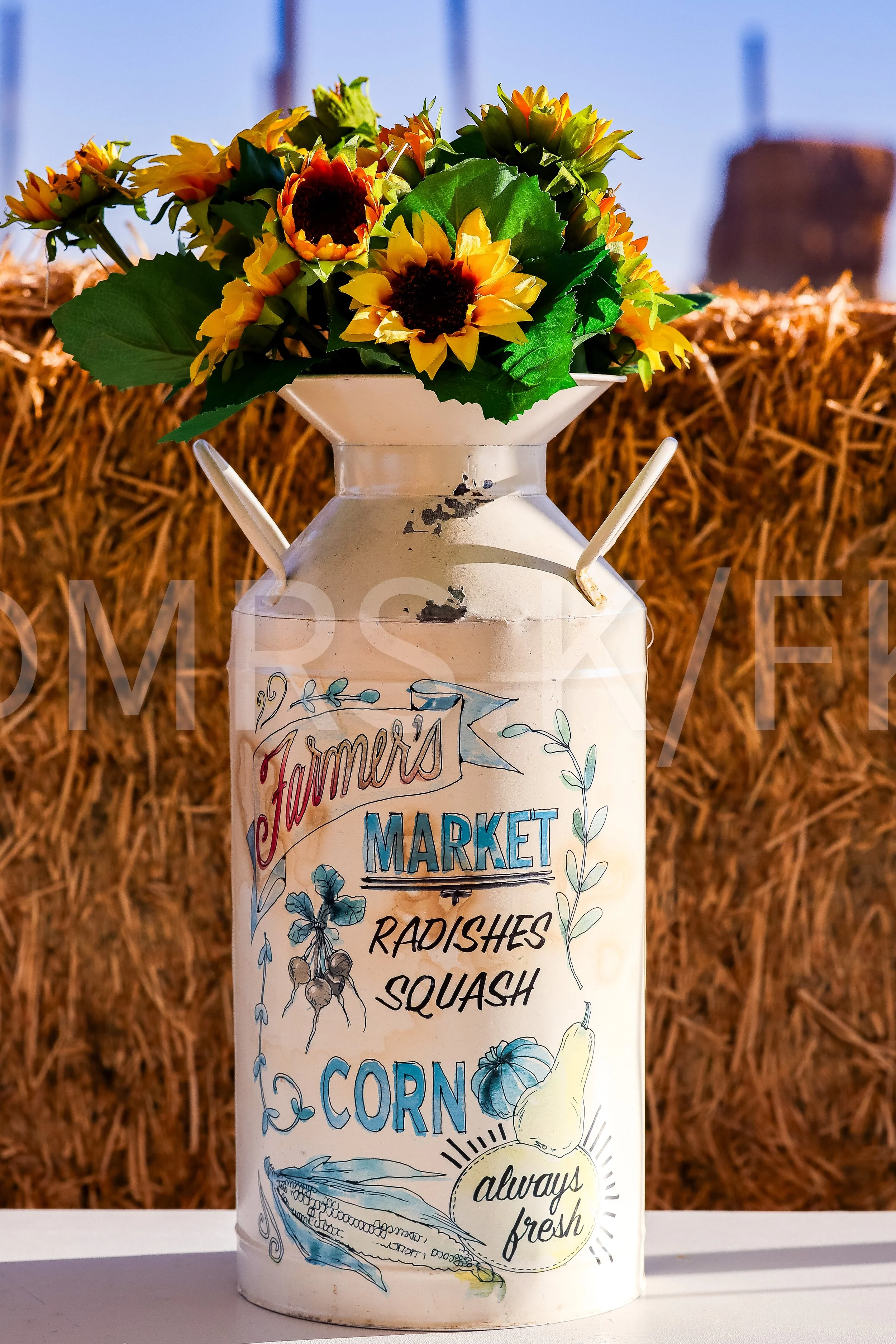 A white container with colorful illustrations and text, filled with a bouquet of yellow and orange sunflowers and greenery, set against a backdrop of hay bales.