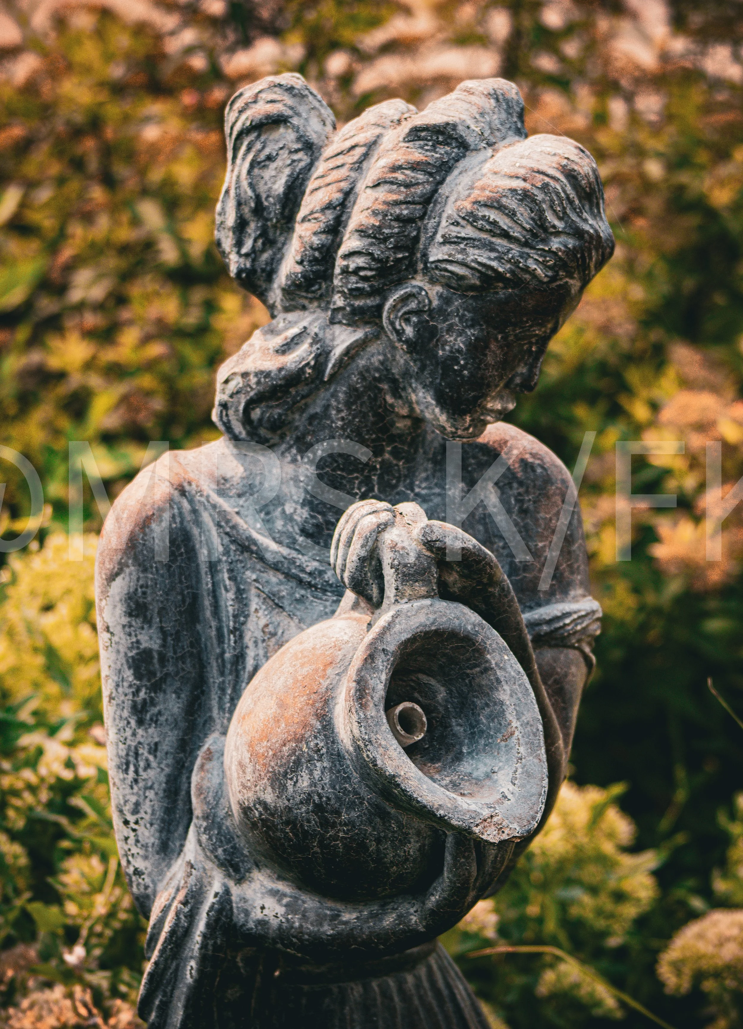 An Original Piece of Photography 1 of 10 to print Custom Fine Art - Close-up of a carved stone angel sculpture holding a circular ring, with a blurred background of green and brown foliage.
