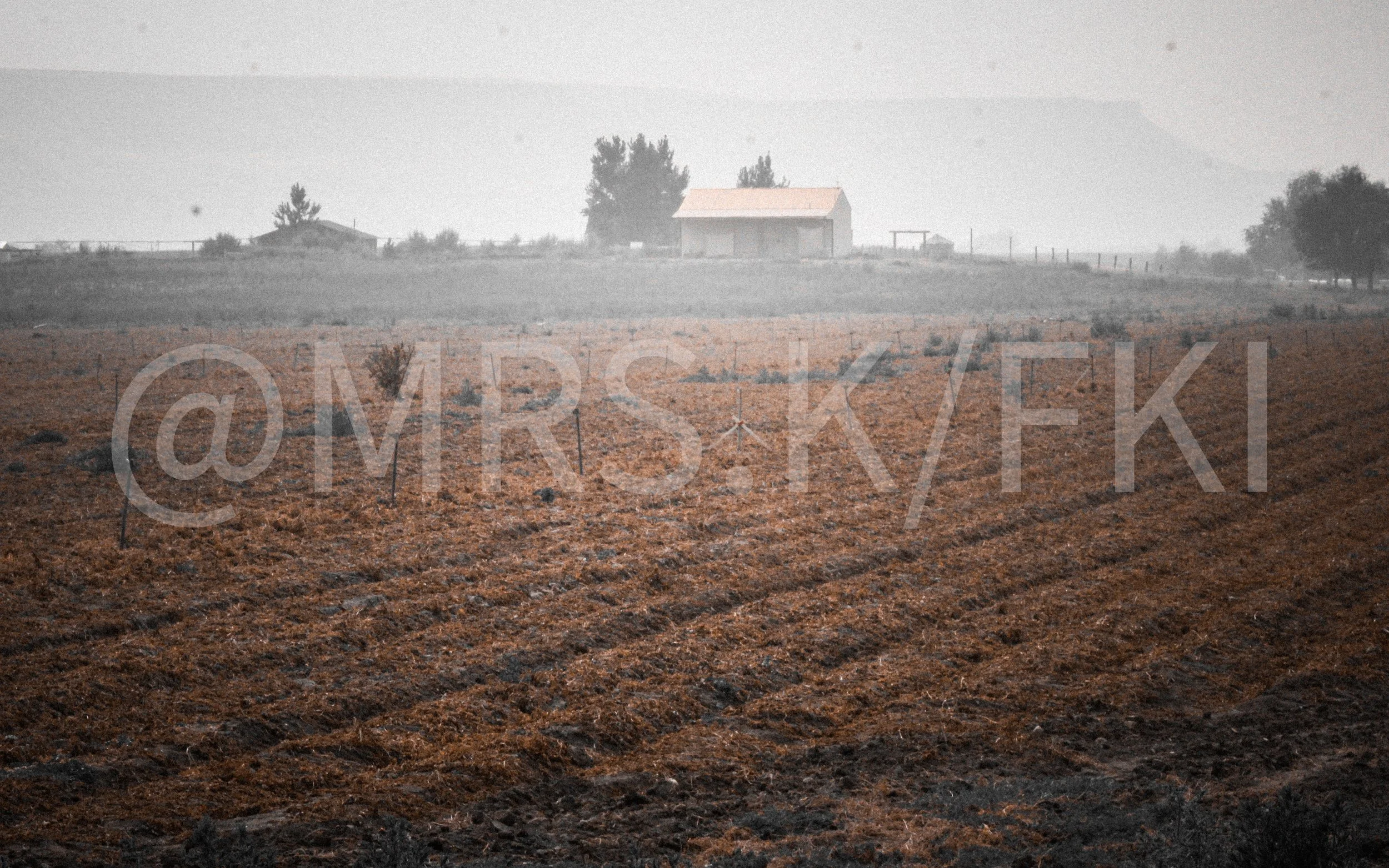 An Original Piece of Photography 1 of 10 to print Custom Fine Art - A rural landscape with a dirt field in the foreground, a small building with a pink roof on a hill, trees, and mountains in the background, under a hazy sky.